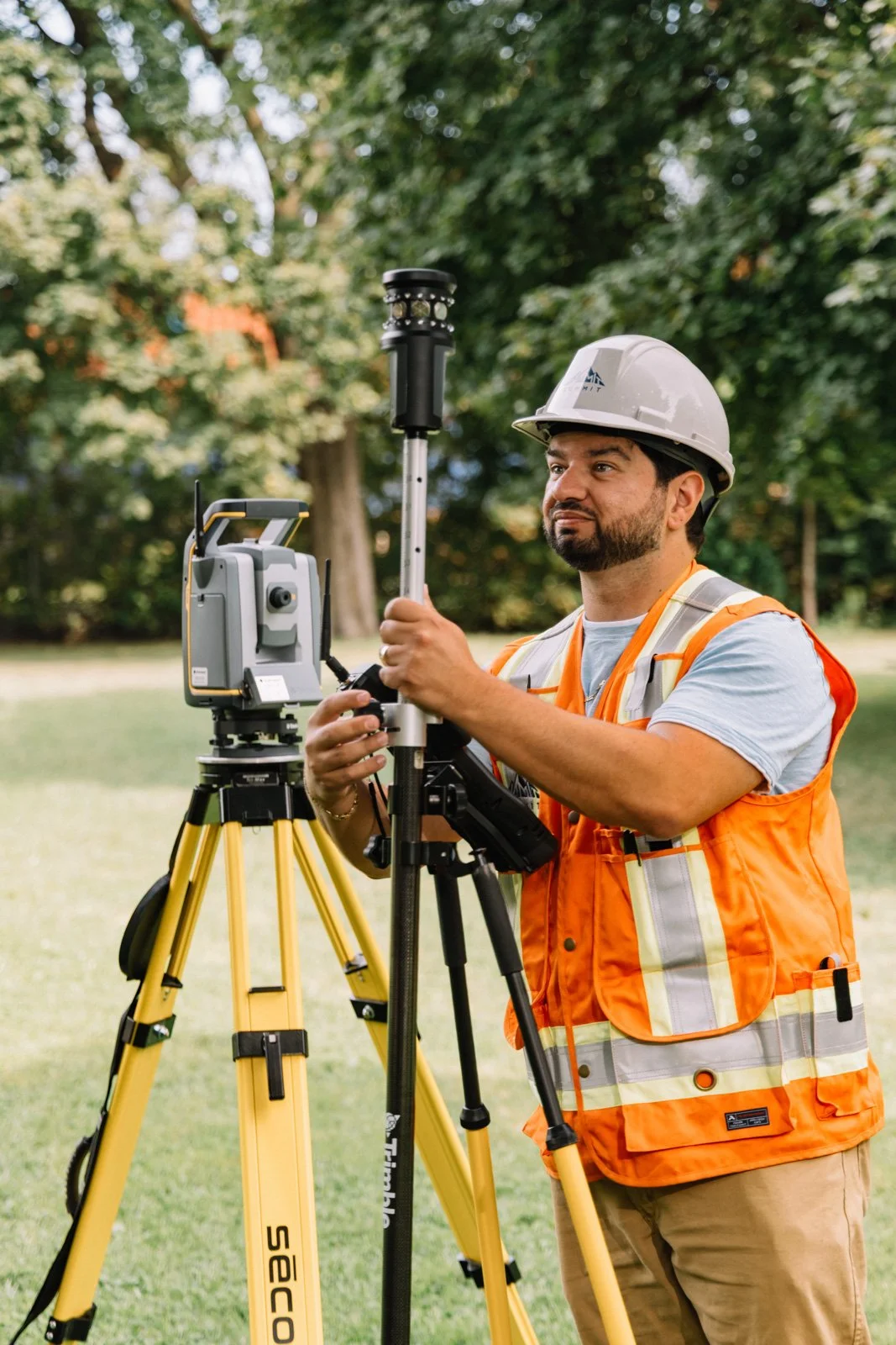A surveyor in safety gear, including a white helmet and orange vest, operating a total station instrument outdoors on a grassy area with trees in the background.