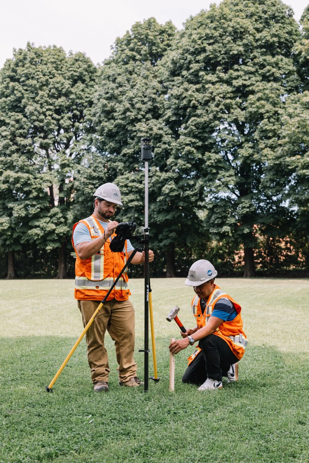 Two construction workers in safety vests and helmets working outdoors with surveying equipment, one standing and the other kneeling with a hammer, on a grassy field with large trees in the background.