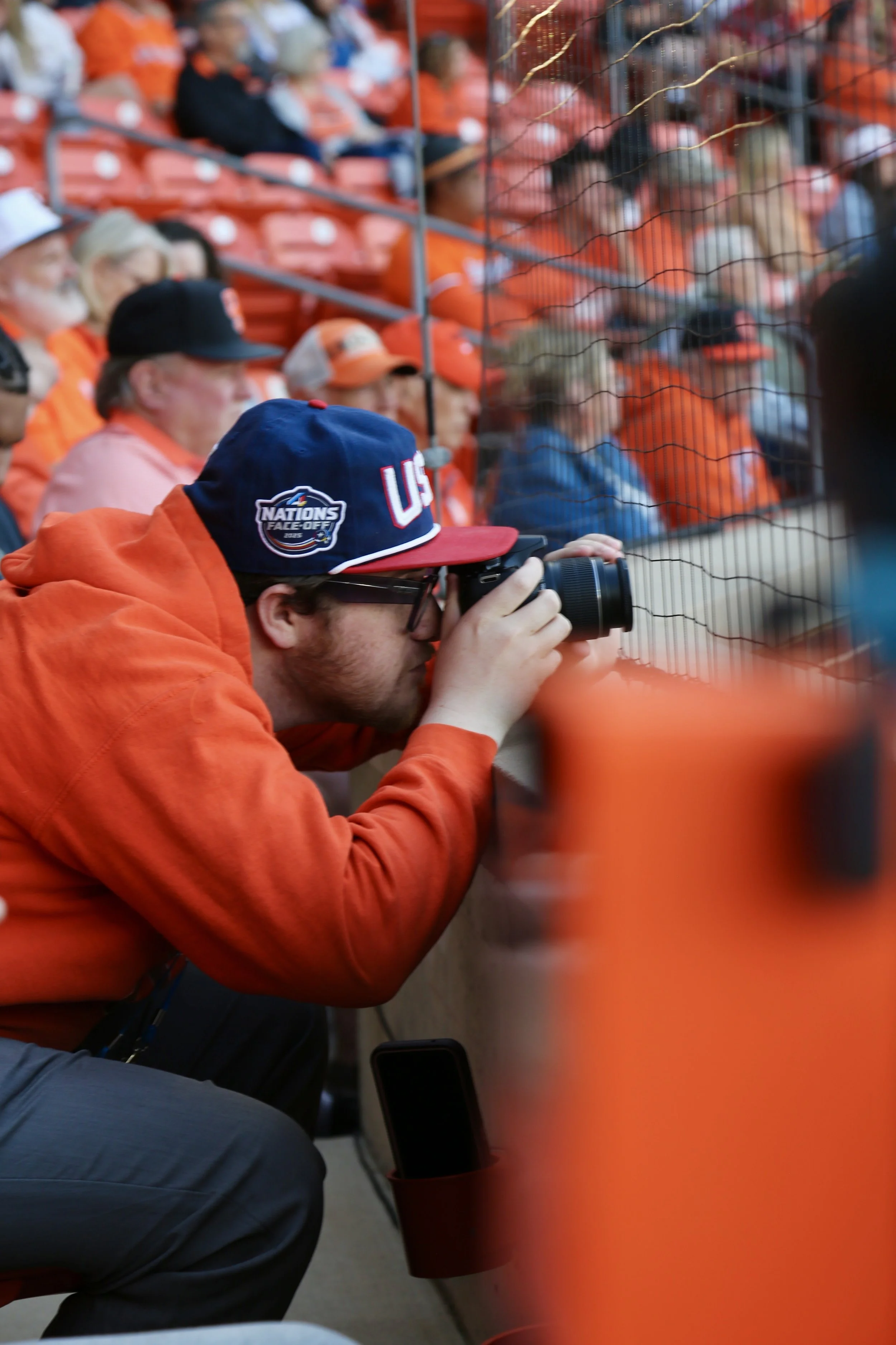 Photographer kneeling and taking a picture at a sports event, all wearing orange, with a metal fence between him and the spectators.