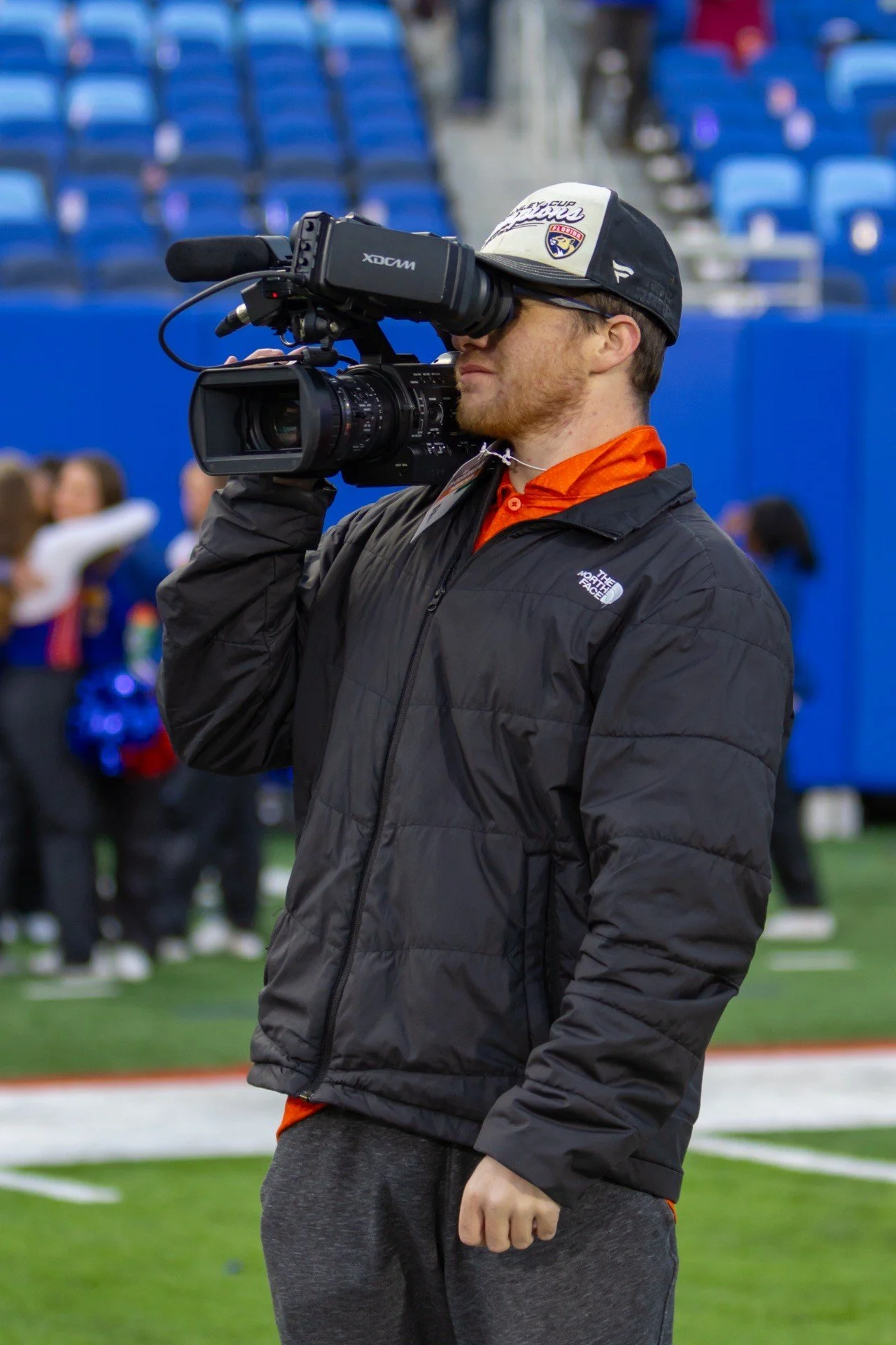 A man standing on a sports field operating a professional video camera, dressed in a black North Face jacket, orange shirt, gray sweatpants, and a black and white baseball cap with logos, with blue stadium seats and people in the background.