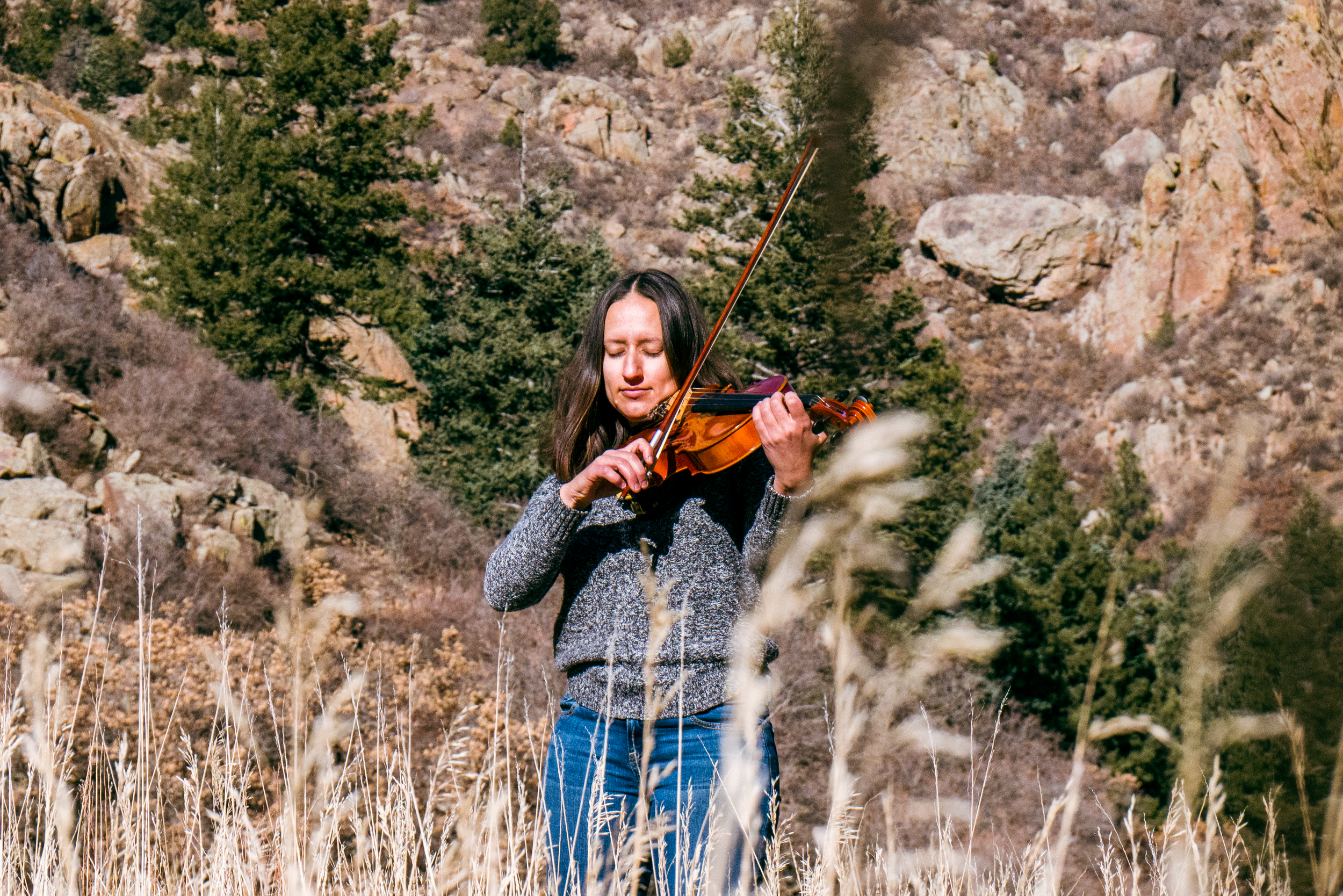 Rebecca MacNamee playing the violin outdoors in a natural, mountainous area with trees and dry grass.
