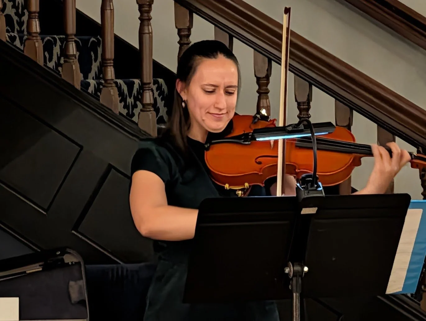 A woman playing the violin indoors, she is standing near a piano, with a music stand in front of her and a staircase with wooden railing behind her.