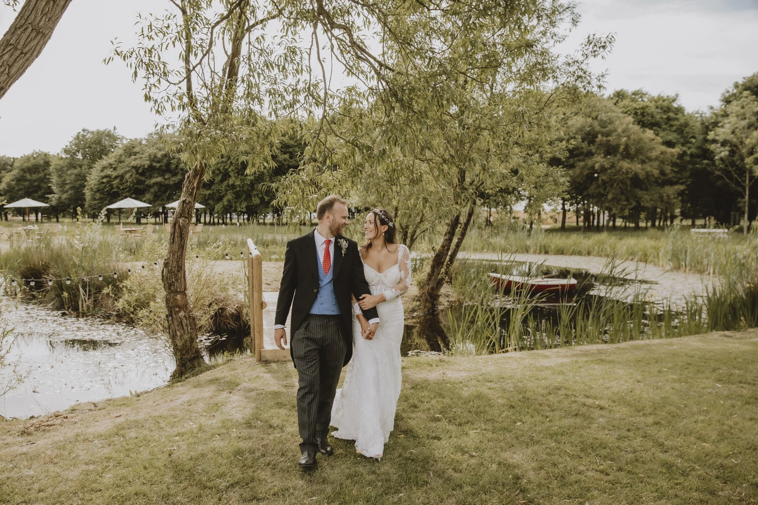 A bride and groom walking hand in hand outdoors near a small pond, surrounded by trees and greenery, with outdoor tables and umbrellas in the background.