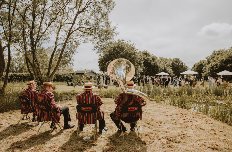 A band wearing matching stripes suits playing instruments outdoors at a garden party with people in the distance near umbrellas and trees on a cloudy day.