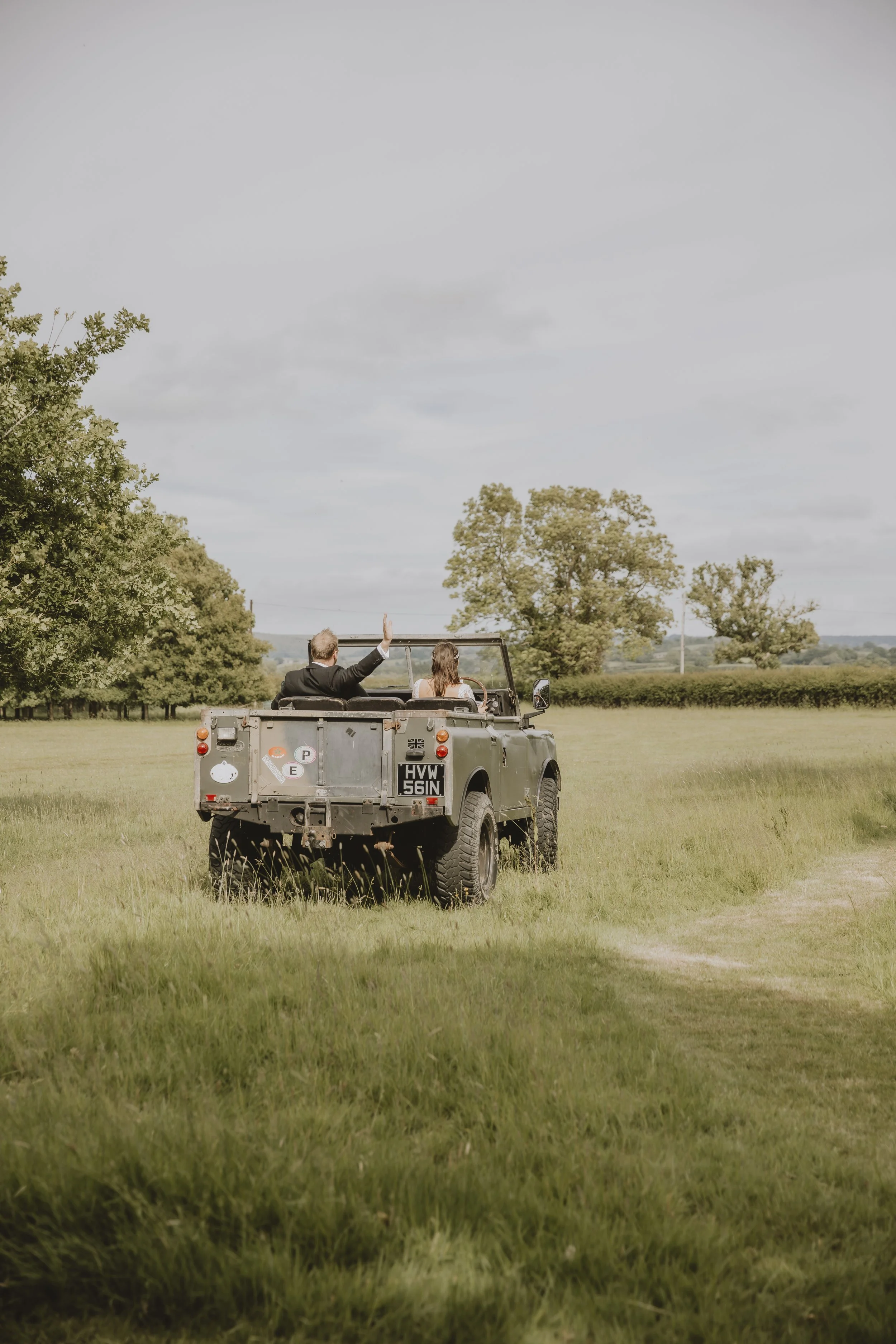 A man and woman in formal attire sitting in the back of a military-style vehicle, waving, while driving through a grassy field with trees in the background.
