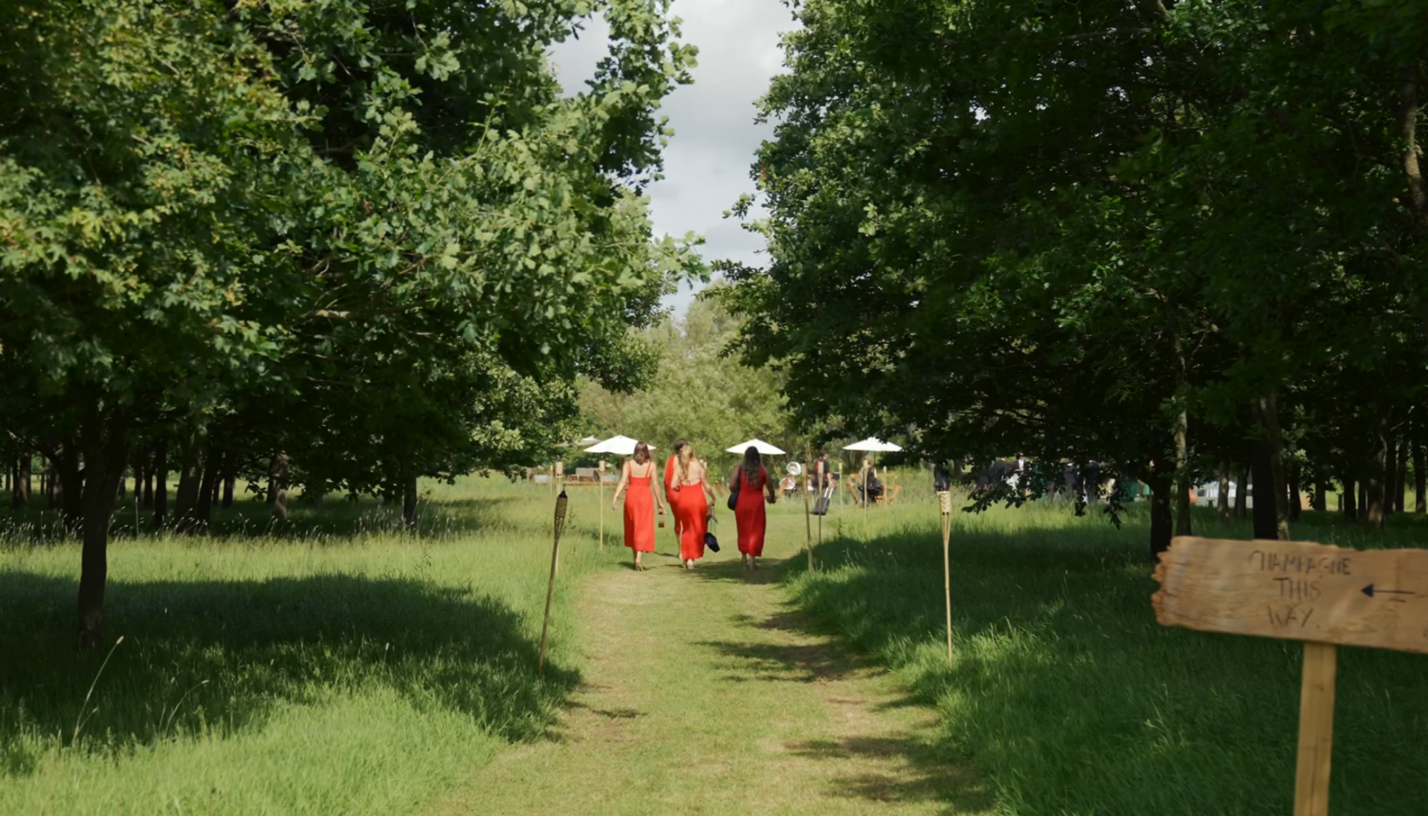 Three women walking along a grassy path in a park with large trees and several white umbrellas in the background.