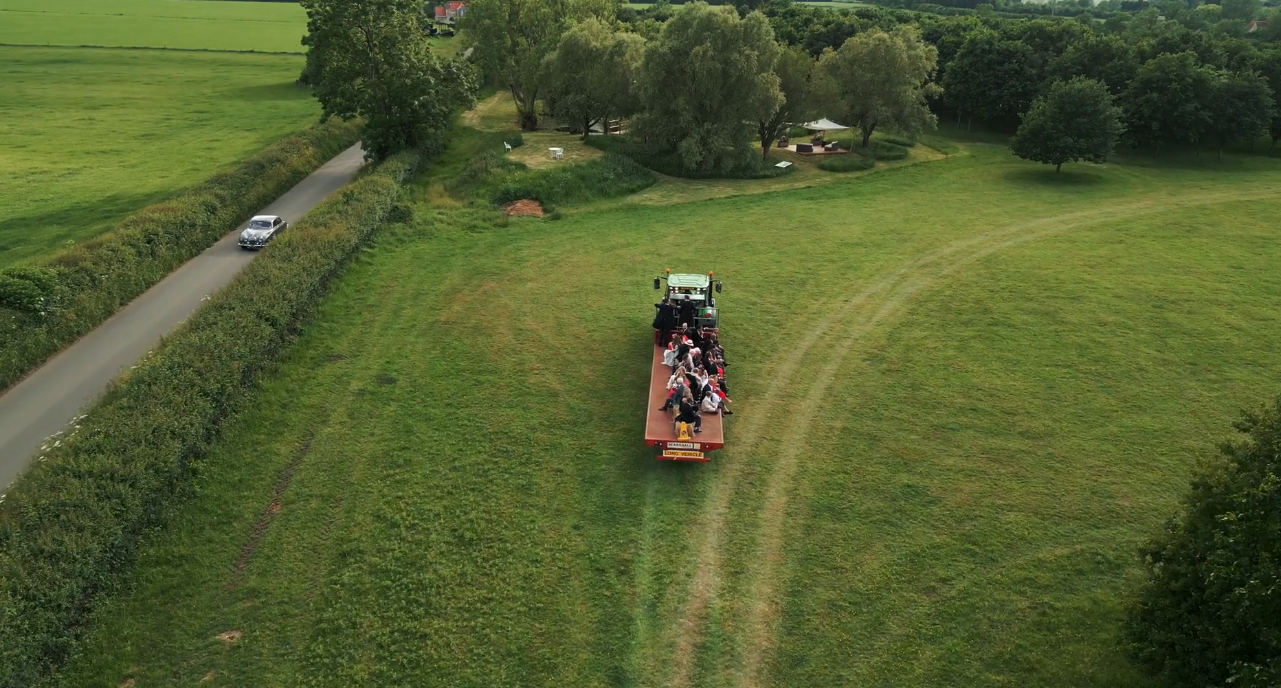 Aerial view of a red flatbed trailer carrying people being pulled by a small green tractor across a grassy field, with a car on a narrow road and trees in the background.