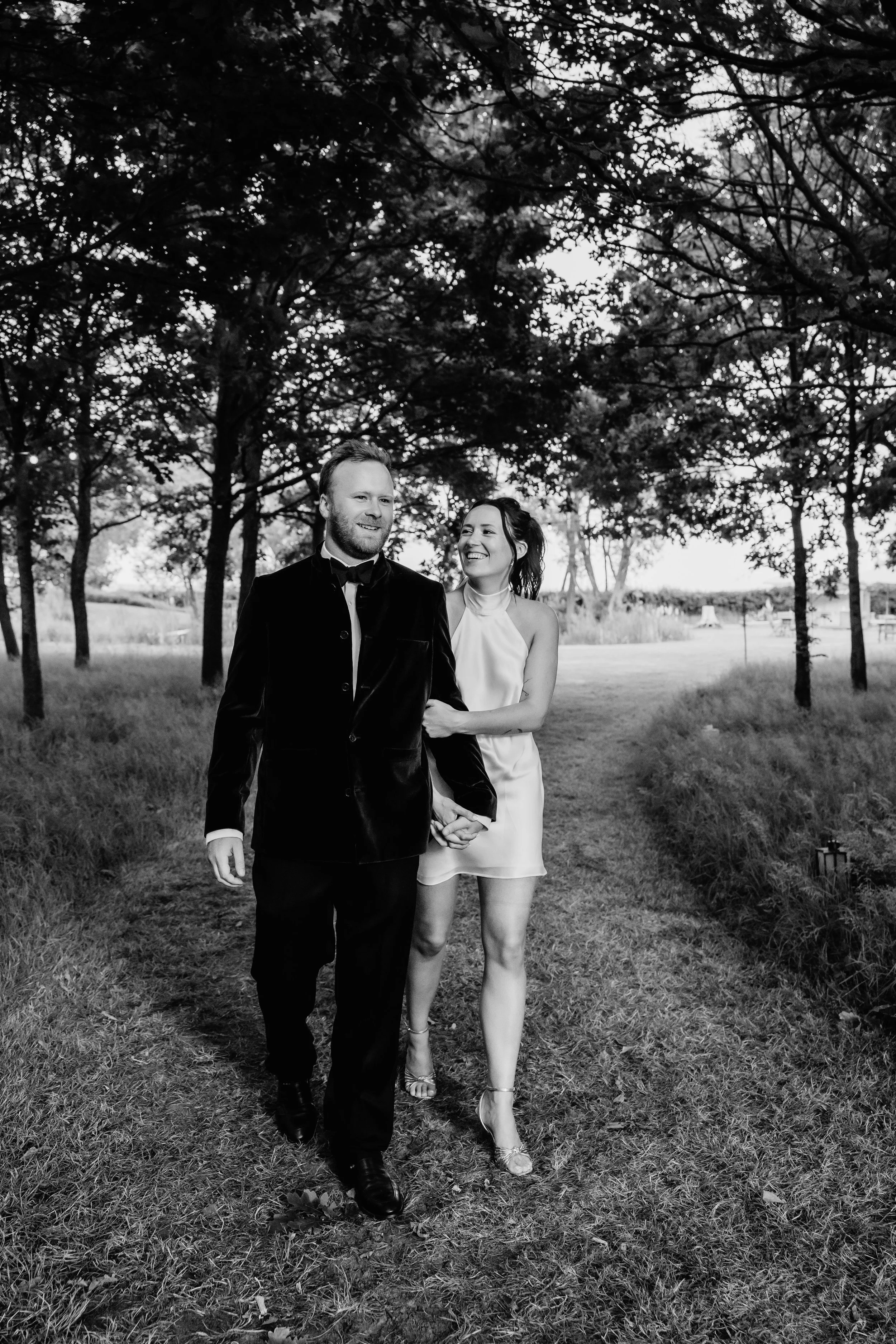 A black and white photo of a man and woman walking outdoors under trees, smiling and holding hands. The man is in a tuxedo and the woman is in a dress and heels.