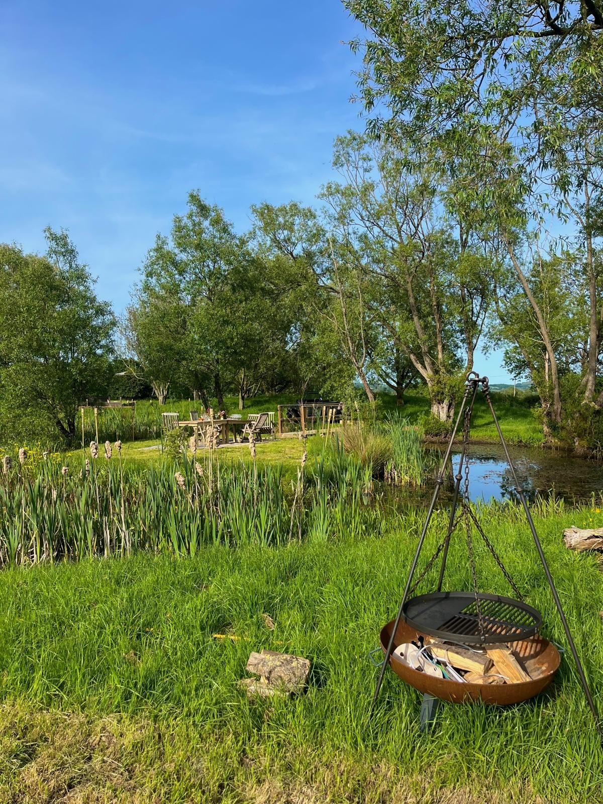 A peaceful outdoor scene with a small pond surrounded by green grass and trees. There is a round table with chairs, a black grill, and a metal stand with a hanging fire pit filled with wood and a grill grate. The sky is clear and blue, indicating a s