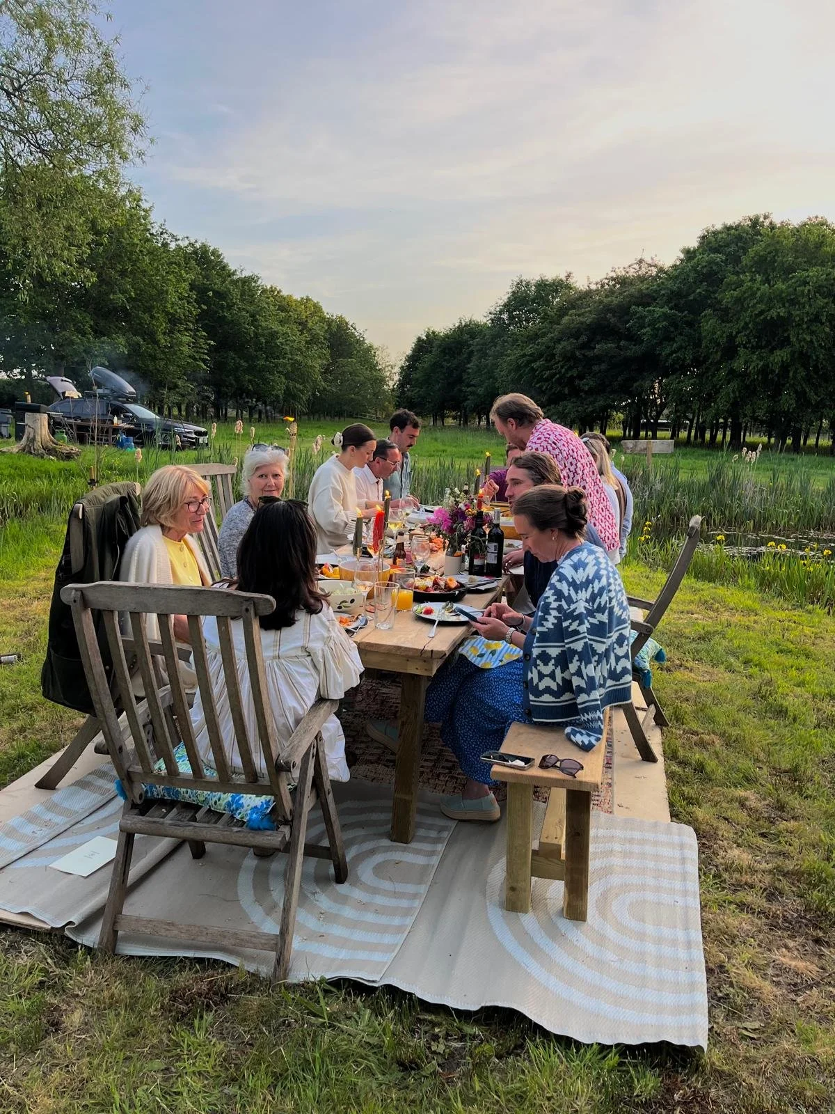 A group of people having dinner outdoors near a pond in a park setting, with trees in the background and a cloudy sky.