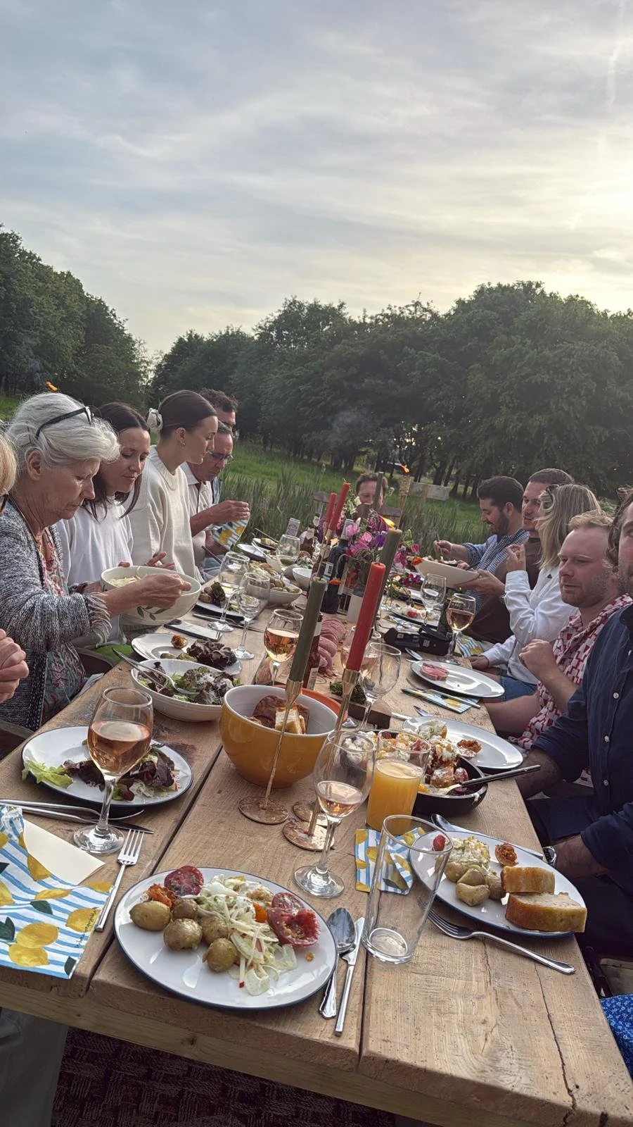 A group of people enjoying an outdoor dinner at a long wooden table with various dishes, glasses of wine, and candles, set in a lush green garden during sunset.