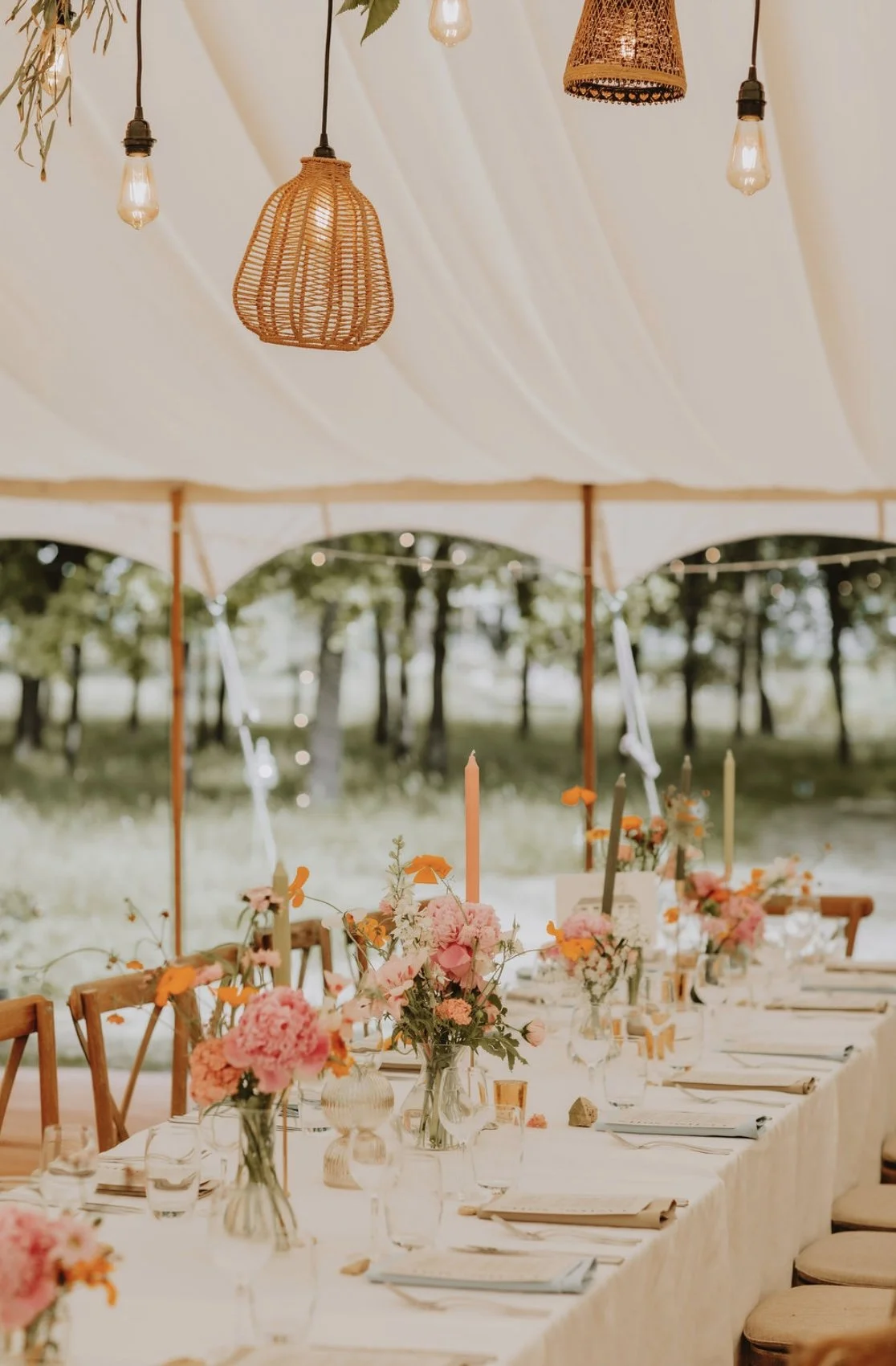 A decorated outdoor dining area under a canopy with hanging wicker and Edison bulb lights, floral centerpieces, candles, and place settings.