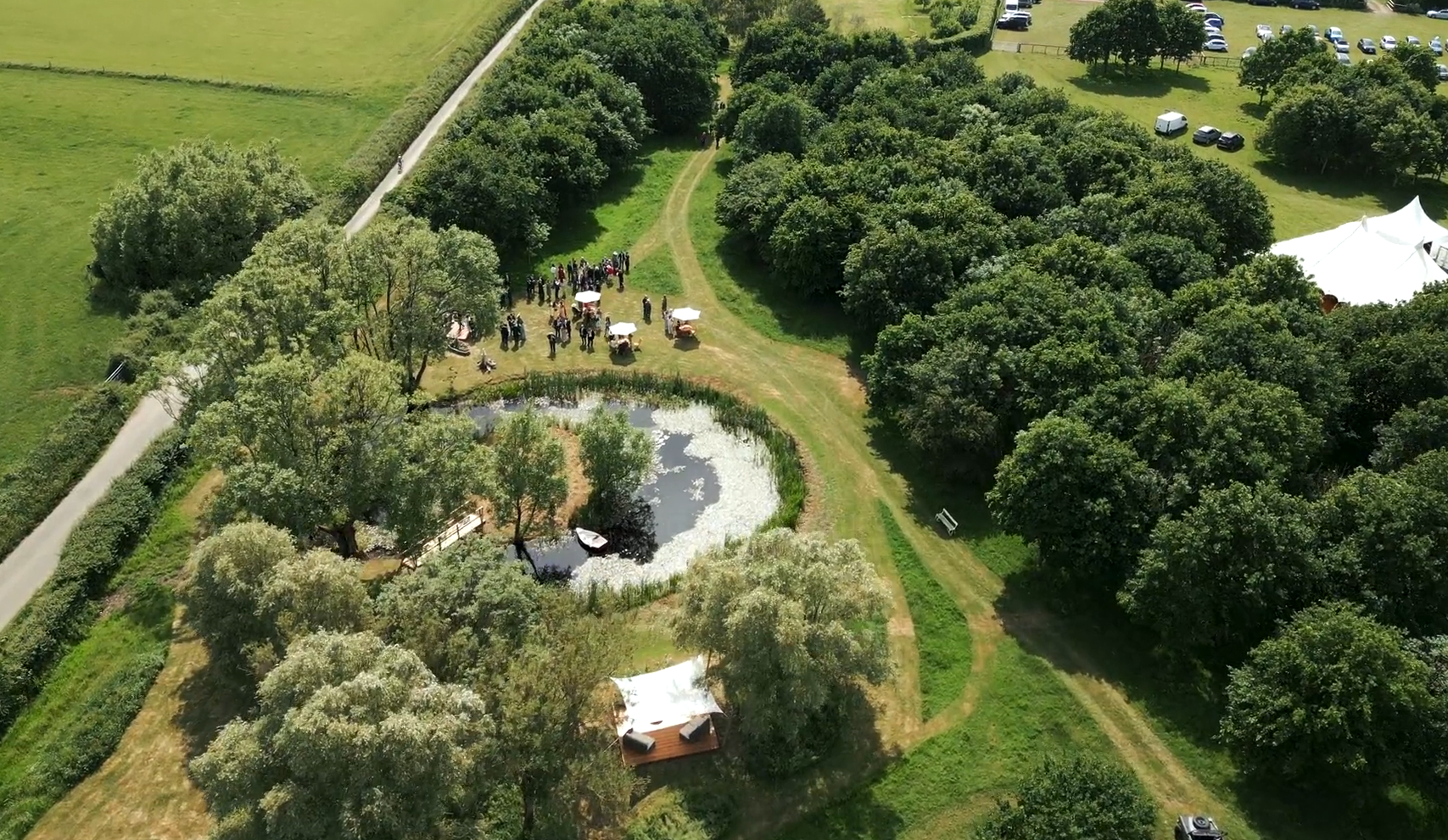 Aerial view of a park with lush green trees, a pond with duckweed, and a gathering of people under umbrellas, near a parking lot and white tent.
