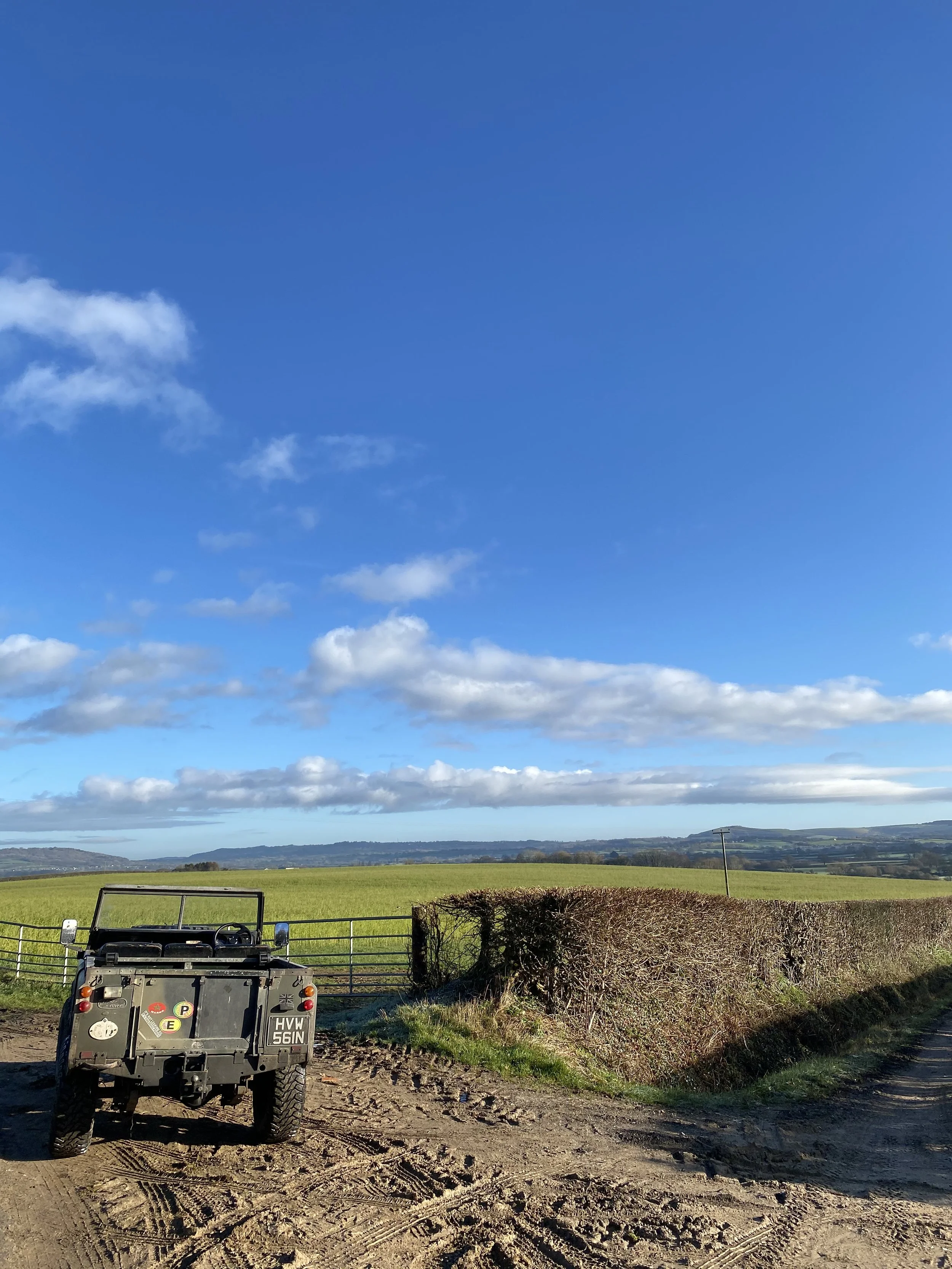 A four-wheeled utility vehicle parked on a dirt road in front of a field of green grass. A hedge and a wire fence are nearby, with rolling hills and a blue sky with scattered clouds in the background.