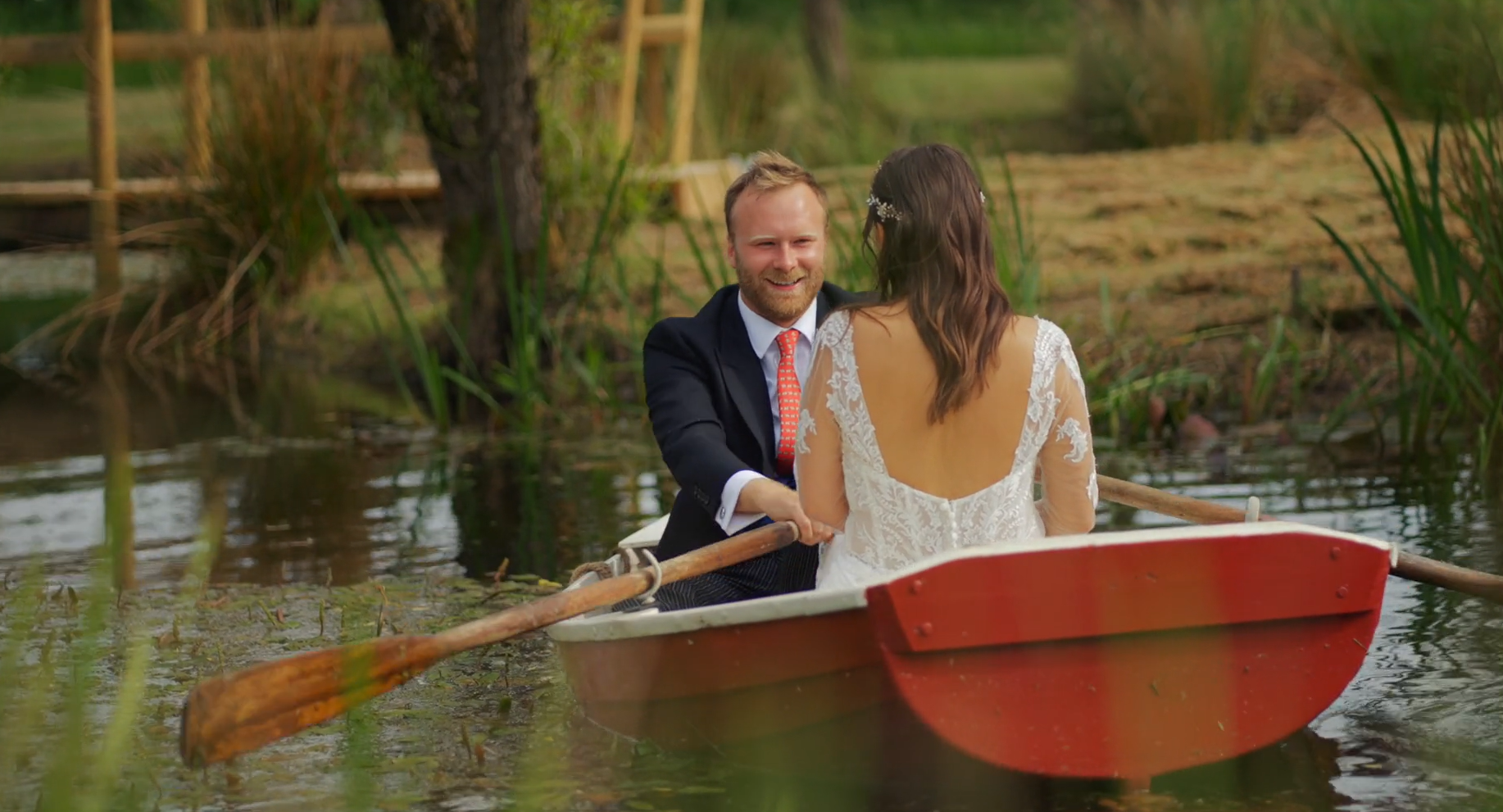 A man and a woman in wedding attire are riding in a small boat on a peaceful pond surrounded by tall grass and trees, with a wooden bridge in the background. The man is smiling at the woman, who is facing away from the camera.