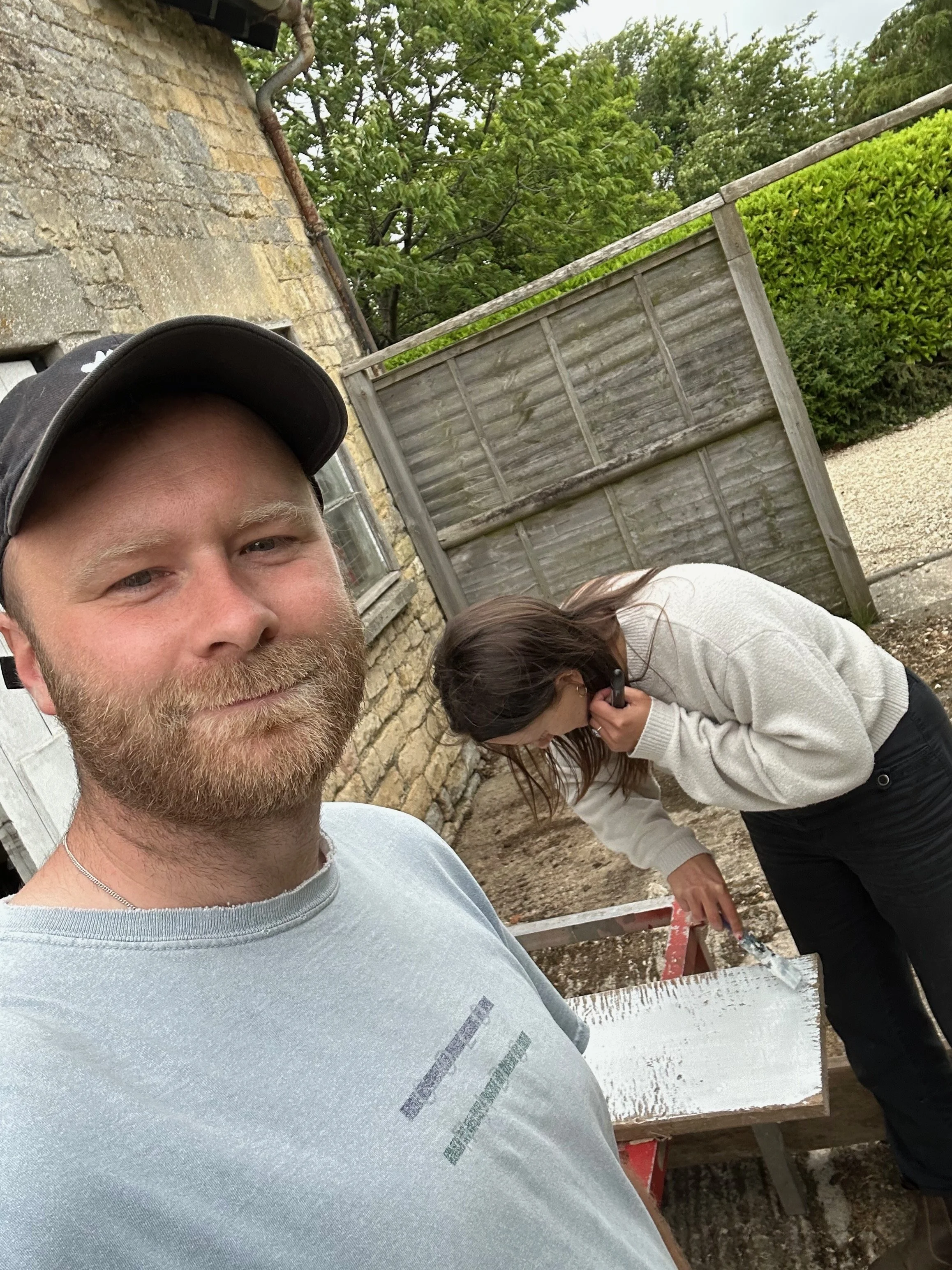 A close-up of a man with a beard and a young woman with glasses, with outdoor construction work: a brick building, wooden fence, and construction tools in the background.