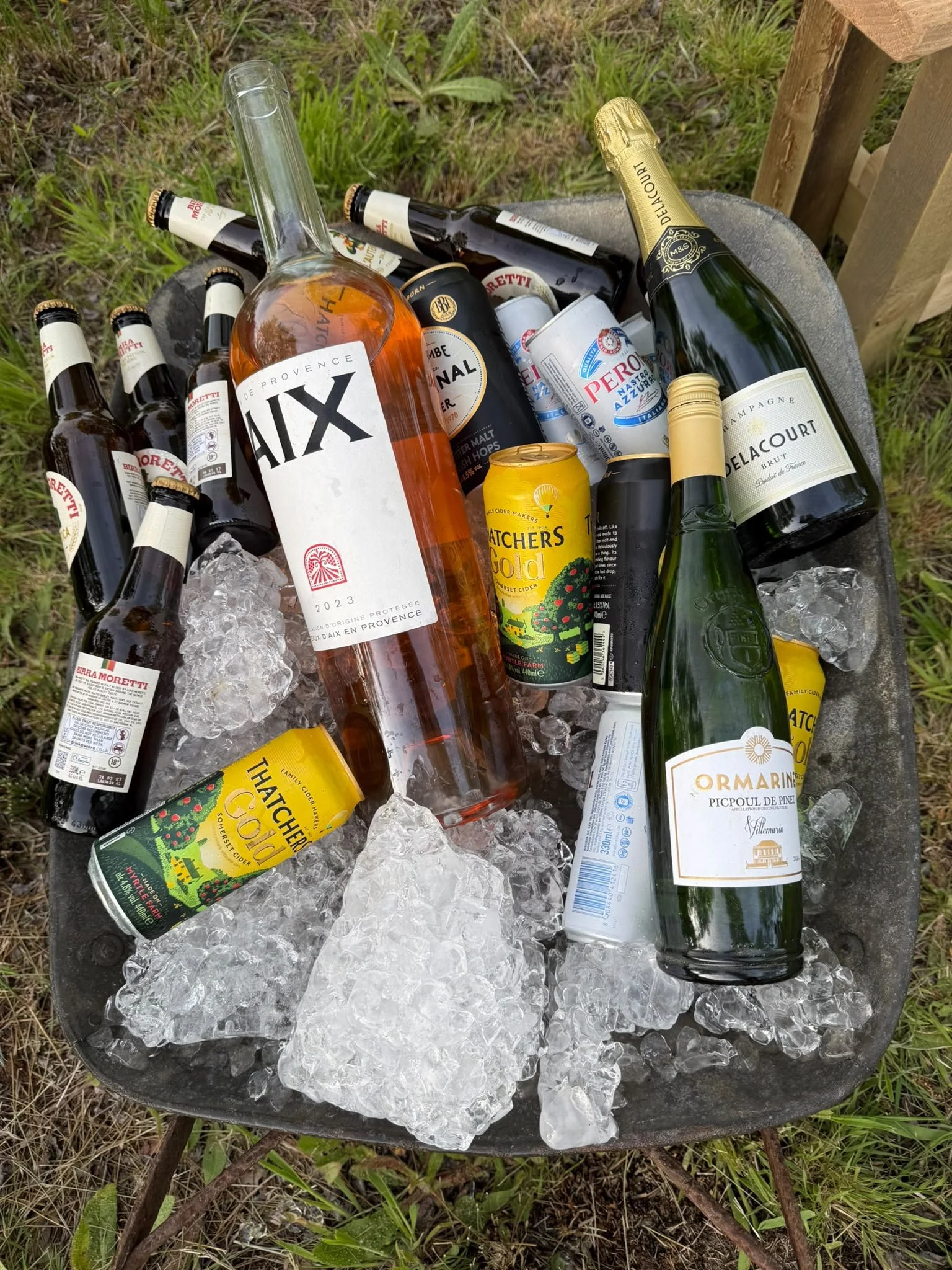 A black tub filled with ice and assorted bottles and cans of alcoholic beverages, including wine, beer, and cider, outdoors on grass.