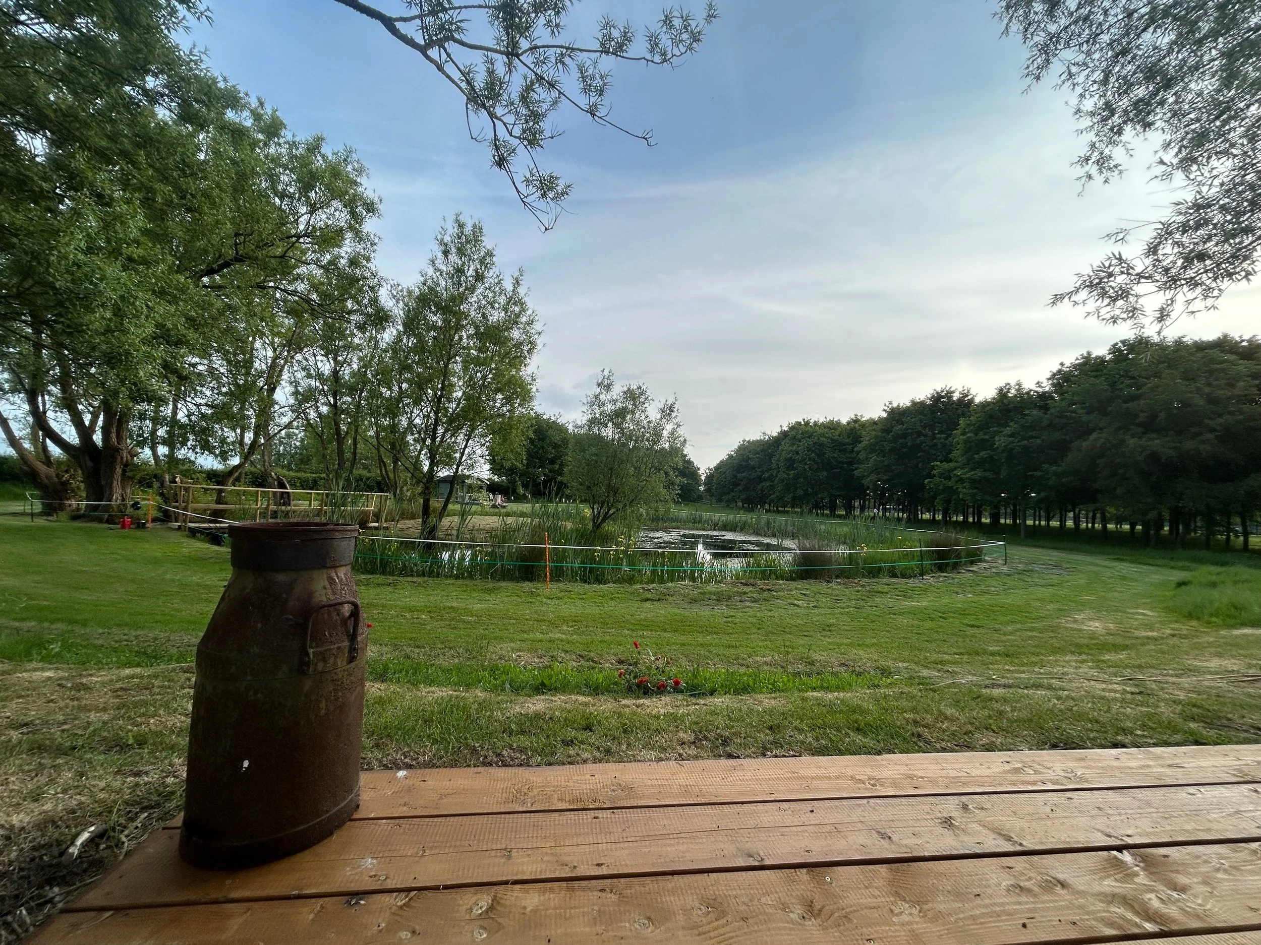 A scenic view of a park with trees, a small pond, a wooden deck, and an old metal barrel in the foreground under a partly cloudy sky.