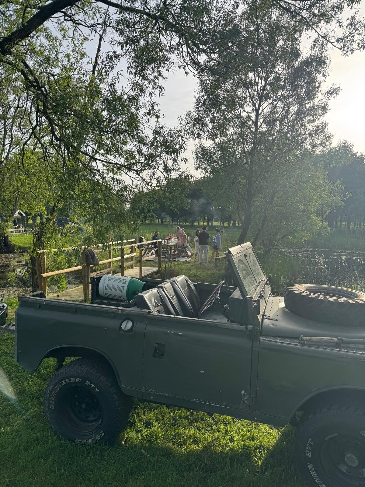 A gray off-road vehicle parked on the grass near a wooden footbridge over a pond, with a group of people gathering at a picnic table in a park surrounded by trees.
