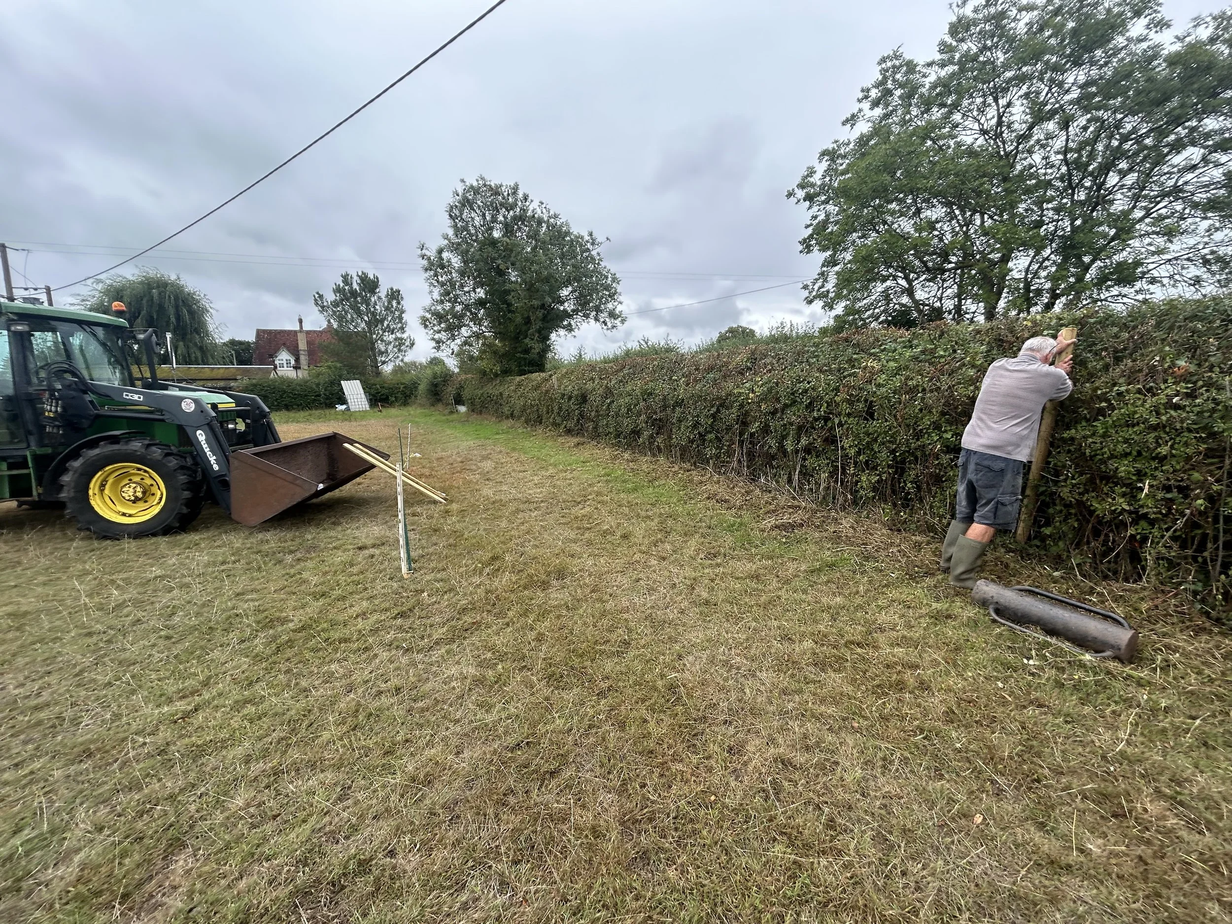 A person trimming a hedge with a pair of hedge shears, standing on the right side of the image. A green tractor with a front loader attachment is on the left side. The scene is outdoors with a cloudy sky, some trees, and a house in the background.