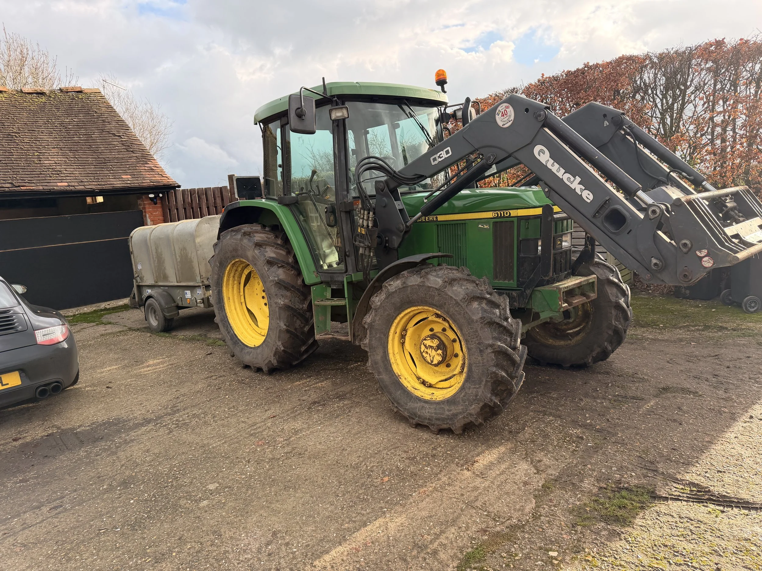 A John Deere tractor with front loader attachment parked on a gravel surface, with older buildings and leafless trees in the background under a partly cloudy sky.