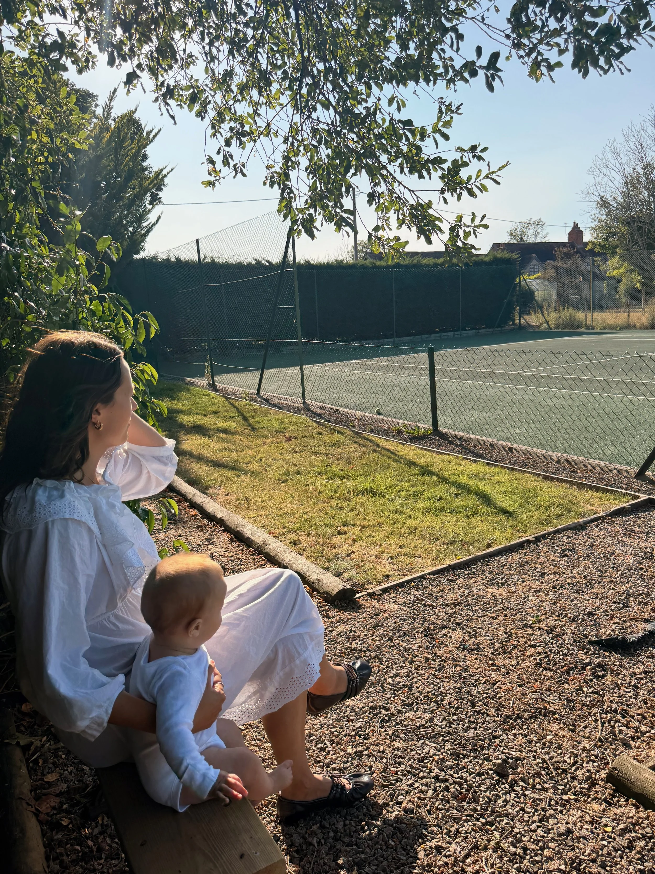 A woman and a young child sitting on a wooden bench outdoors, with a tennis court and trees in the background, under a clear sky.