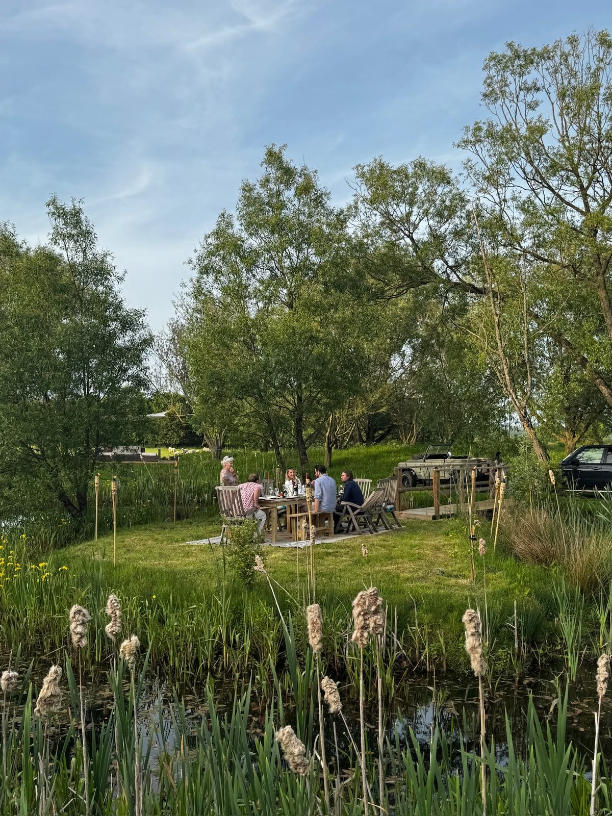Group of six people sitting around a wooden table in a lush garden, surrounded by trees and a pond with tall grasses in the foreground, on a clear day.