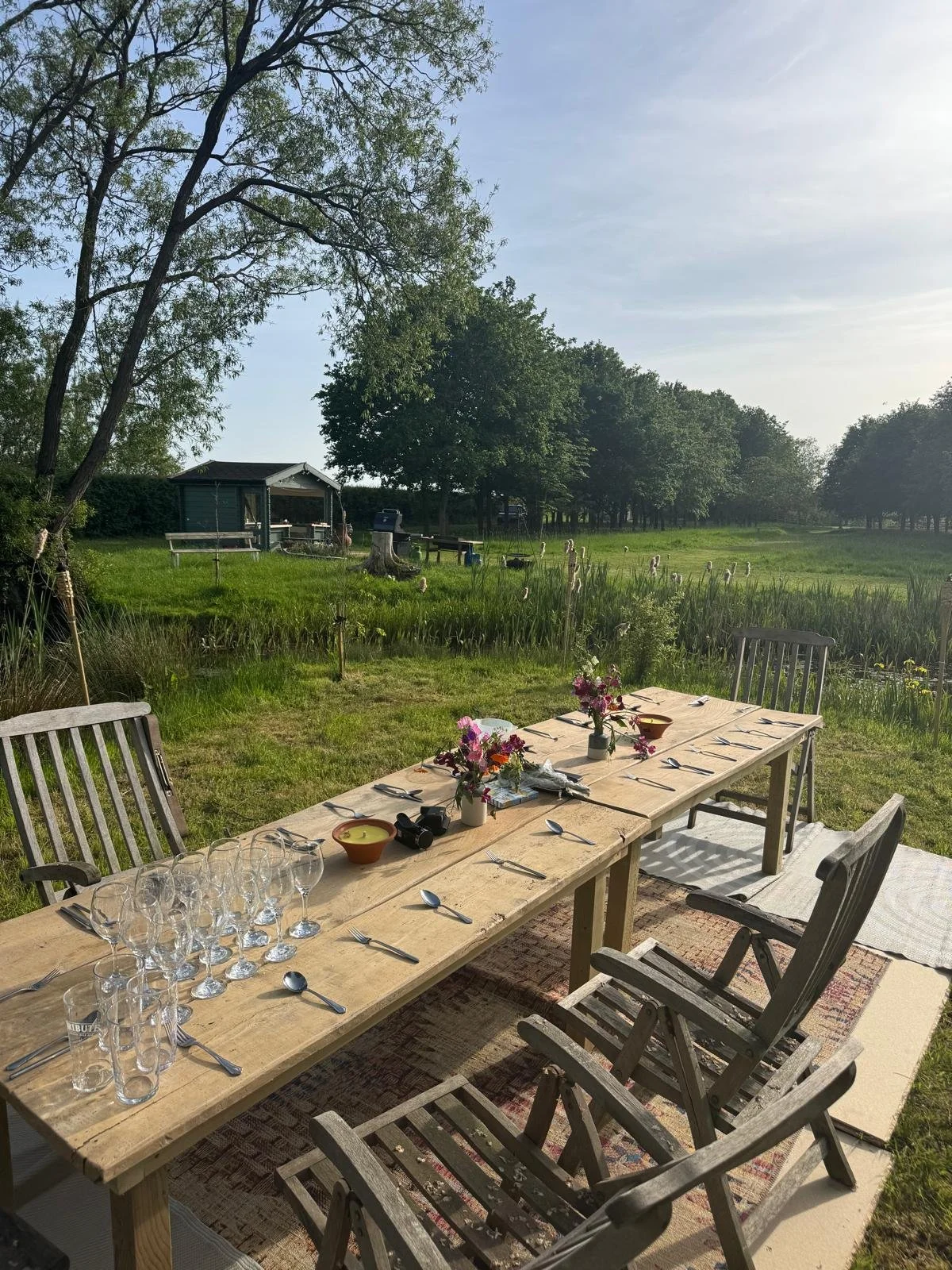 A long outdoor dining table set with glasses, silverware, and floral centerpieces, on a patio with a grassy yard, trees, and a small shed in the background during daylight.