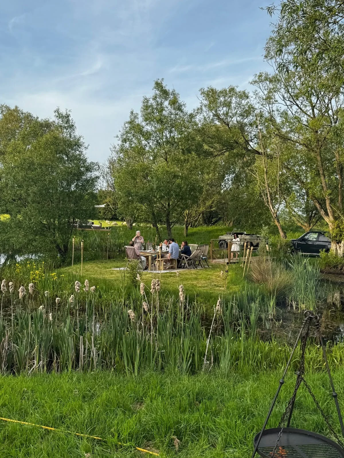 People enjoying a meal outdoors by a pond surrounded by trees and tall grass, with cars and a person standing near a picnic table in the background.