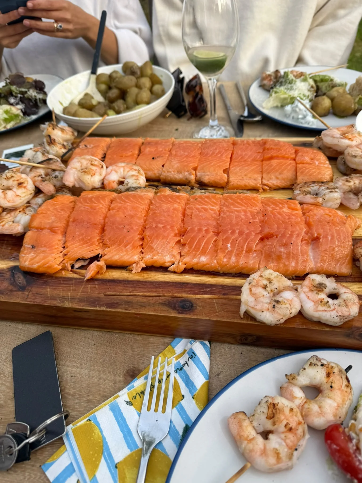 A wooden table with grilled shrimp skewers, salmon fillets, and a white plate containing baked shrimp, with a fork, a floral napkin, and a black lighter in the foreground, and a glass of white wine with a lime wedge, bowls of green olives and salad, 