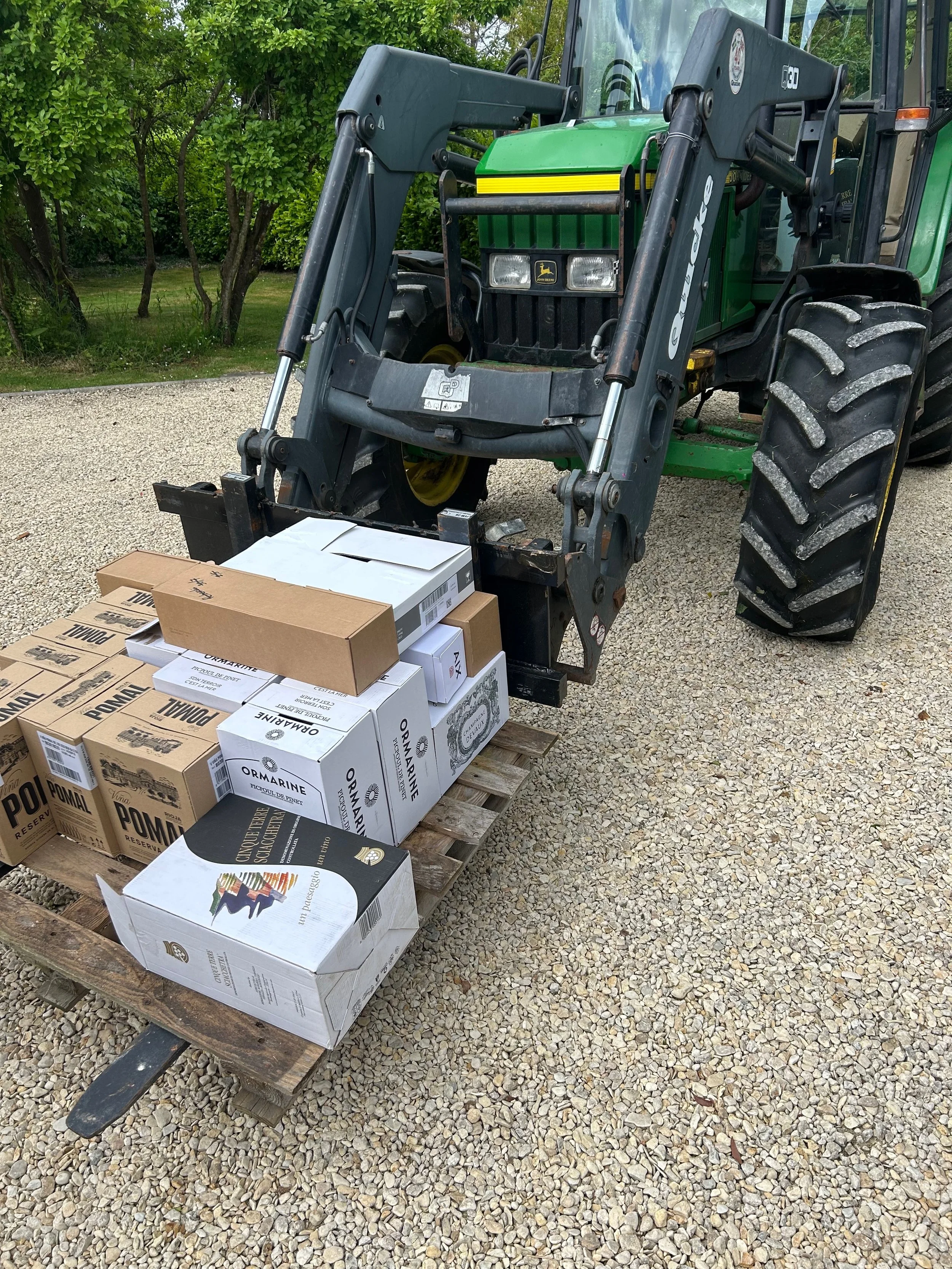A green and black tractor with a front loader attachment is parked on a gravel surface. The tractor has large tires, and there are several boxed items on a wooden pallet in front of it, including white and brown boxes labeled with various text.