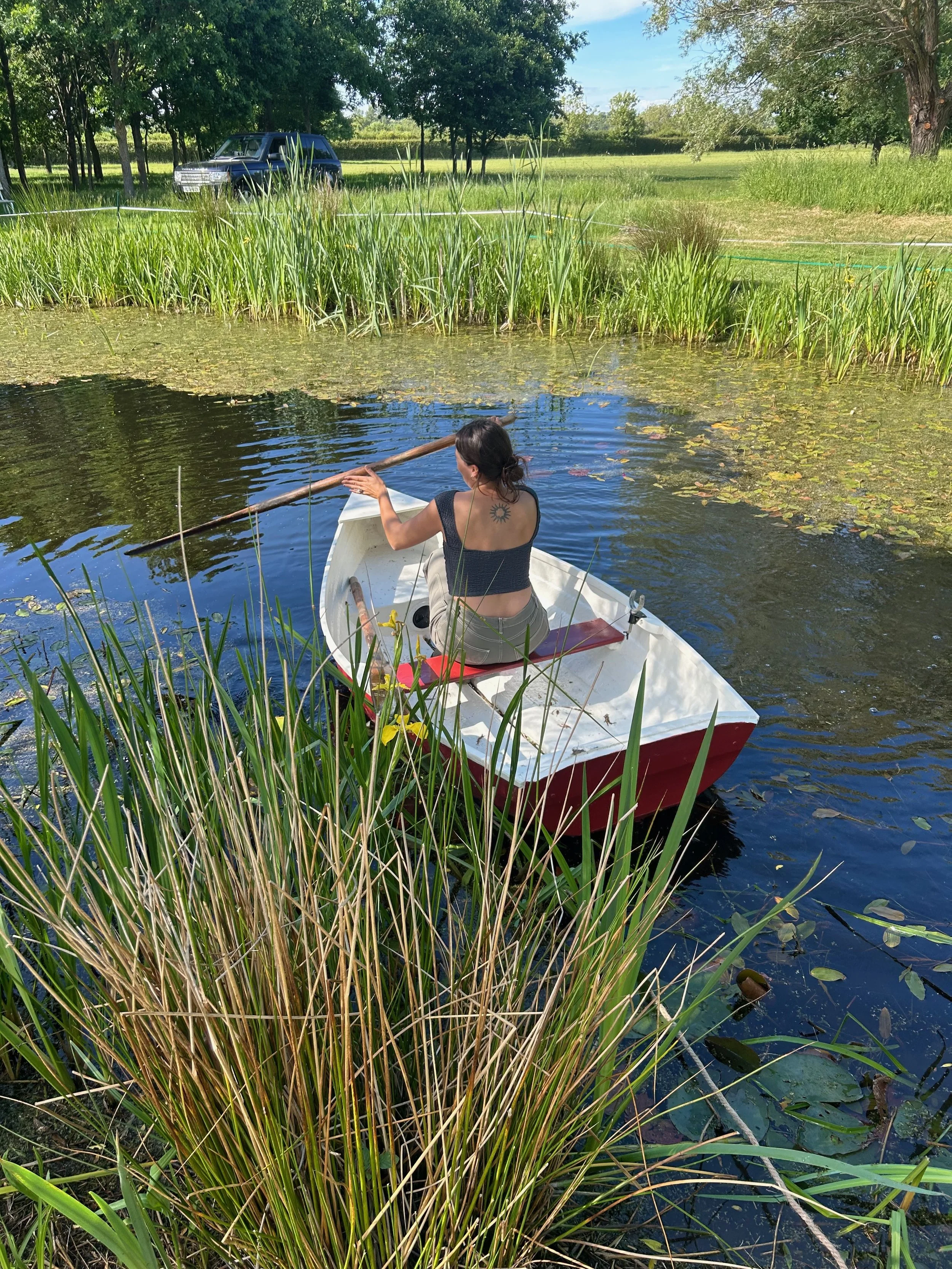 A woman with a sun tattoo on her back sits in a small white boat on a pond covered with lily pads, surrounded by tall grass and trees on a sunny day.
