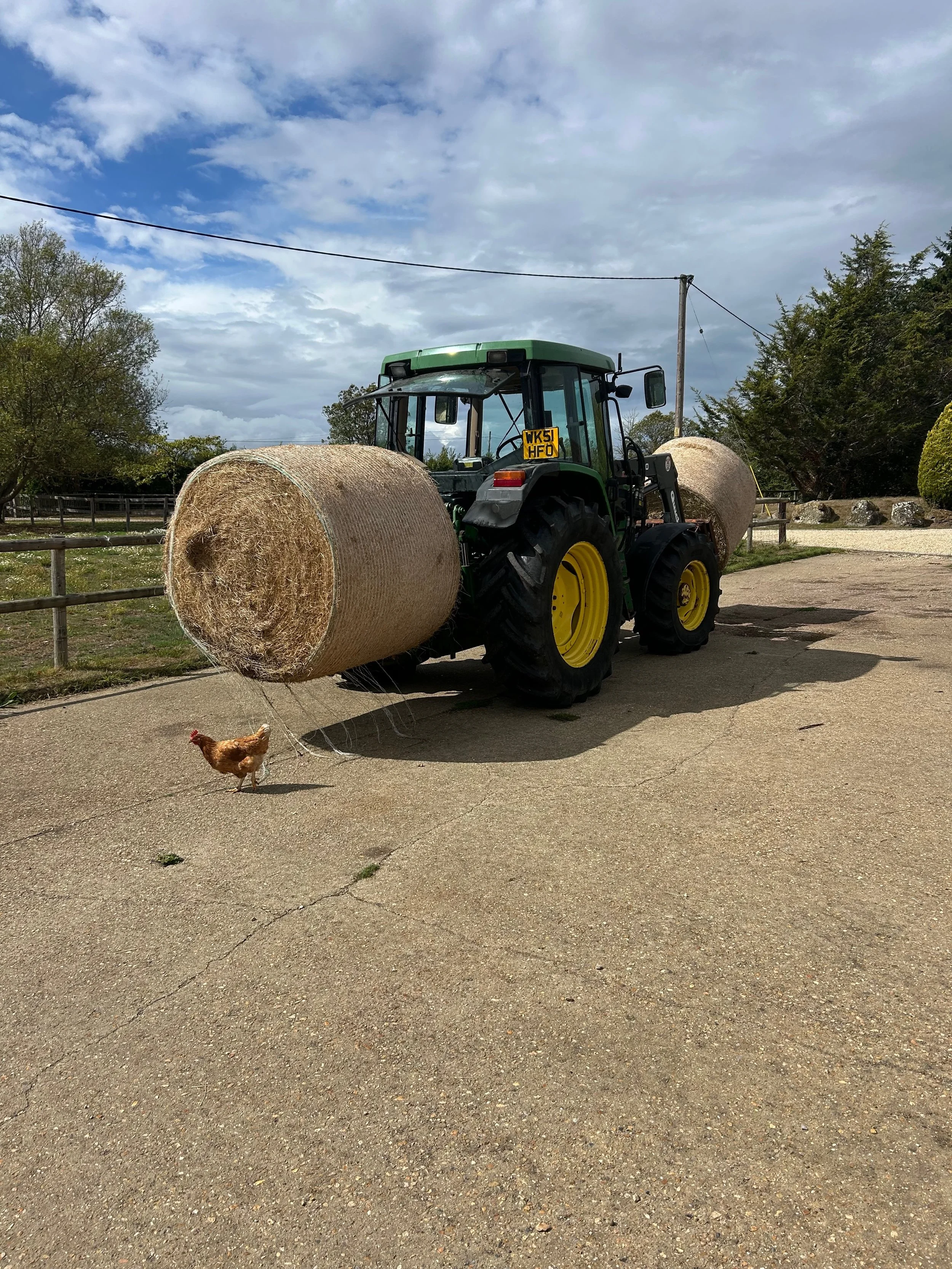 A green tractor with large black tires and a yellow license plate parked on a paved surface with two large hay bales behind it. A chicken is in the foreground near the tractor. The background features trees, a wooden fence, utility poles, and a partl