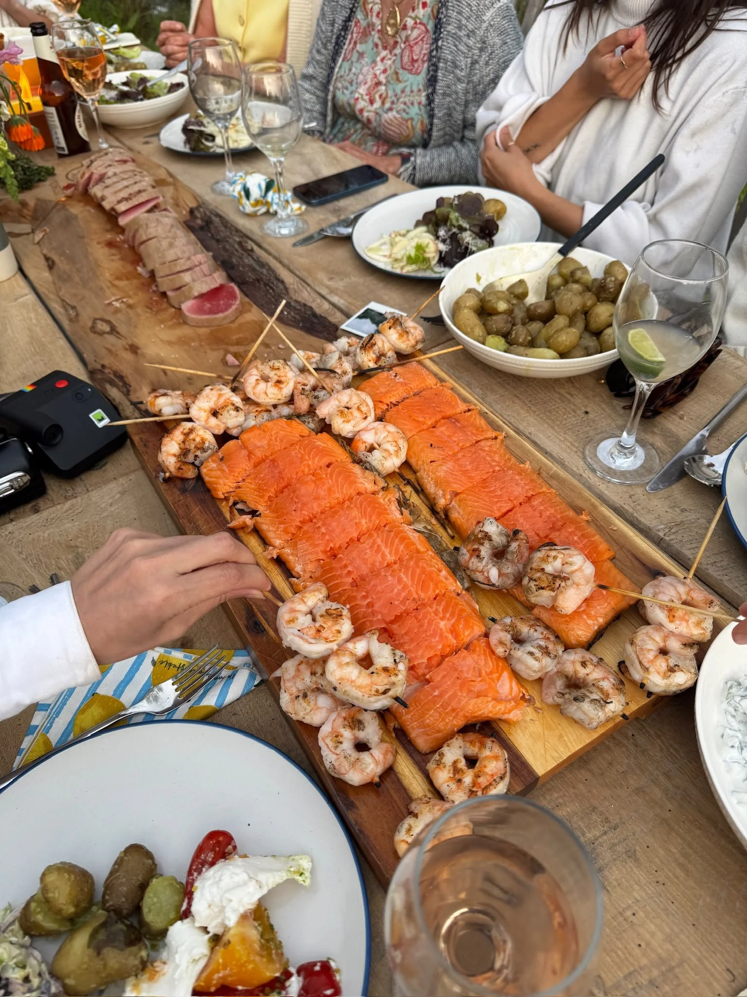 A wooden table with various dishes, including smoked salmon, shrimp skewers, a bowl of baby potatoes, and a plate of greens with dressing. Several empty wine glasses, a smartphone, and a small camera are also on the table. Several people are sitting 