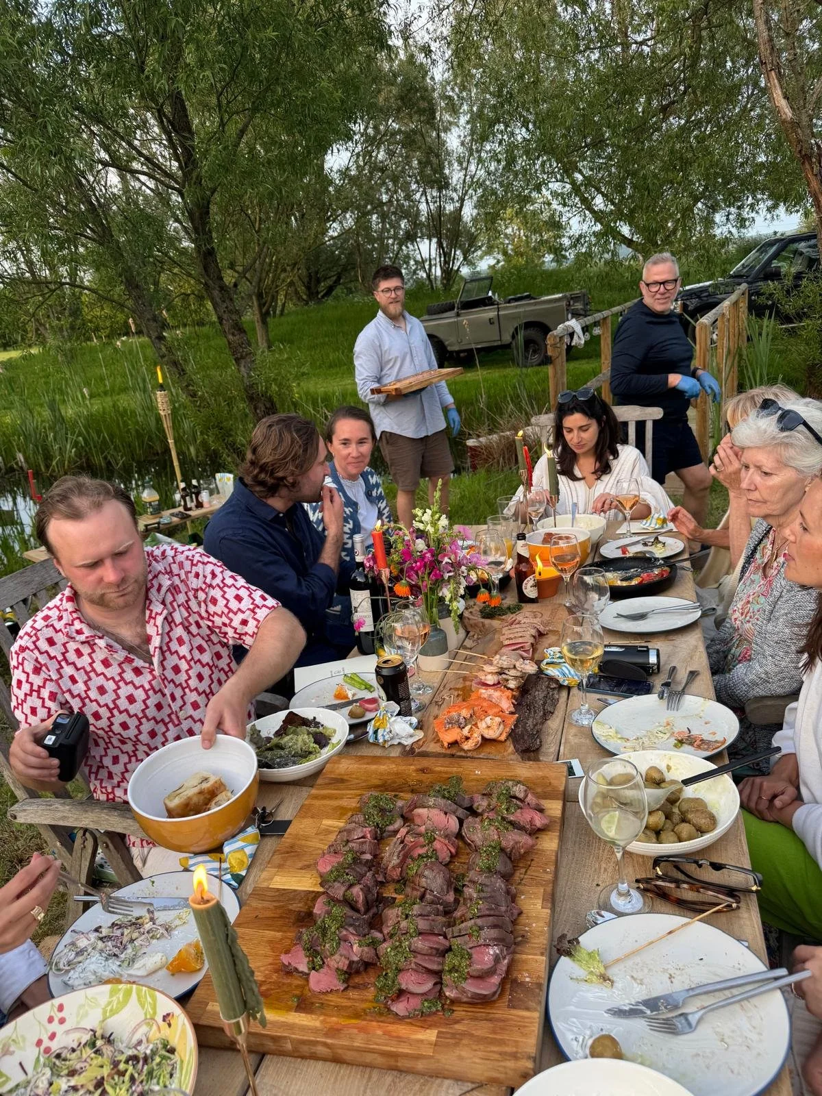 A group of people enjoying an outdoor dinner by a lake, with food, drinks, and flowers on the table, while some others are serving food in the background.