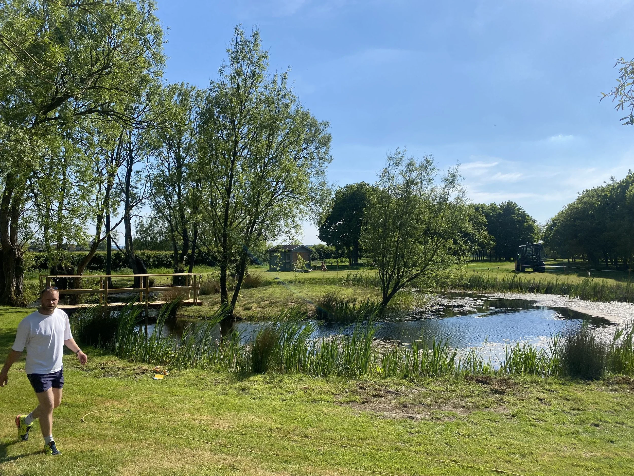 A man walking near a pond with tall grass, trees, and a small wooden structure in the background on a bright, sunny day.