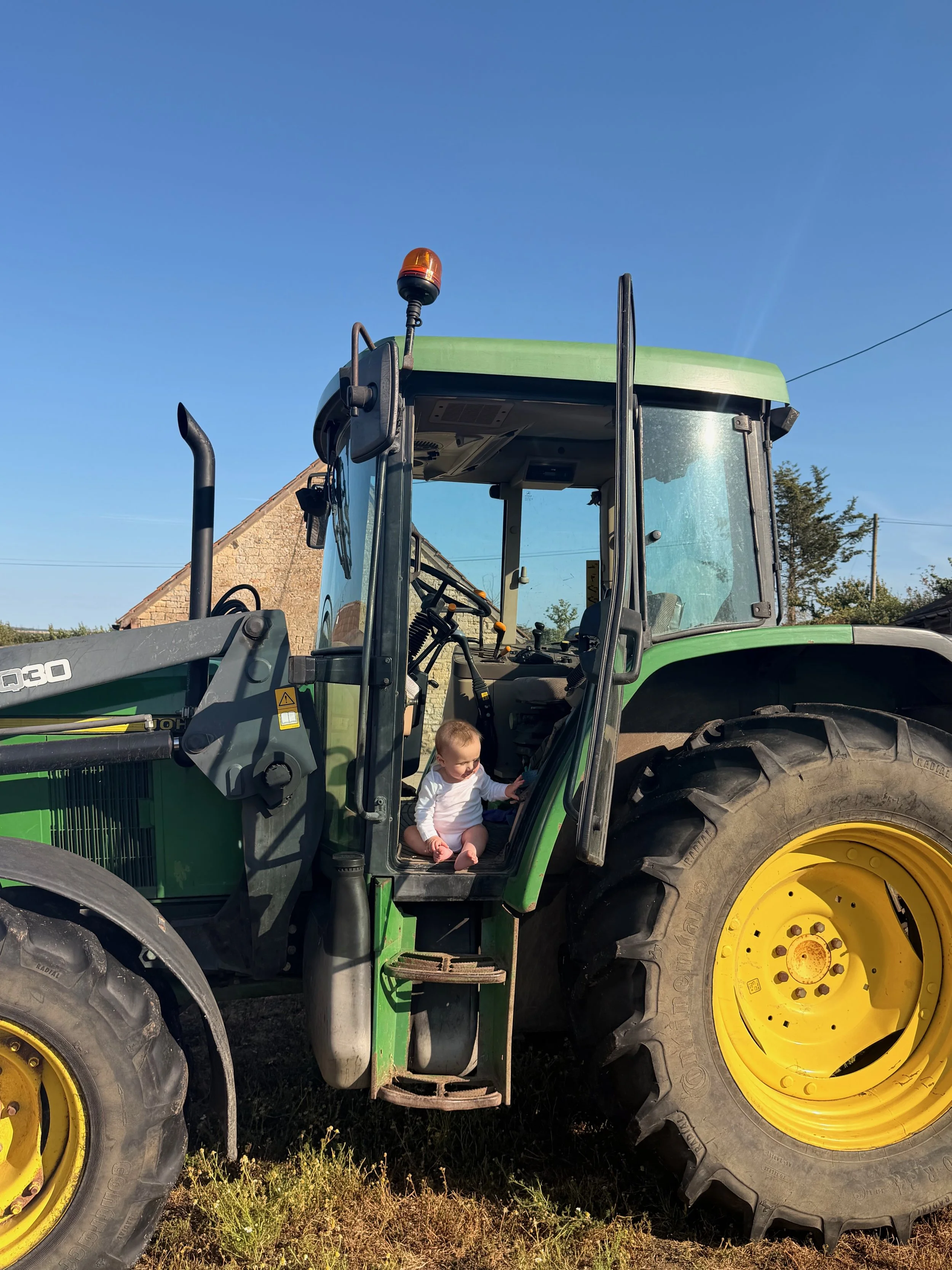 A young child sitting inside the cabin of a large green and yellow tractor on a farm, with a brick building and power lines in the background under a clear blue sky.