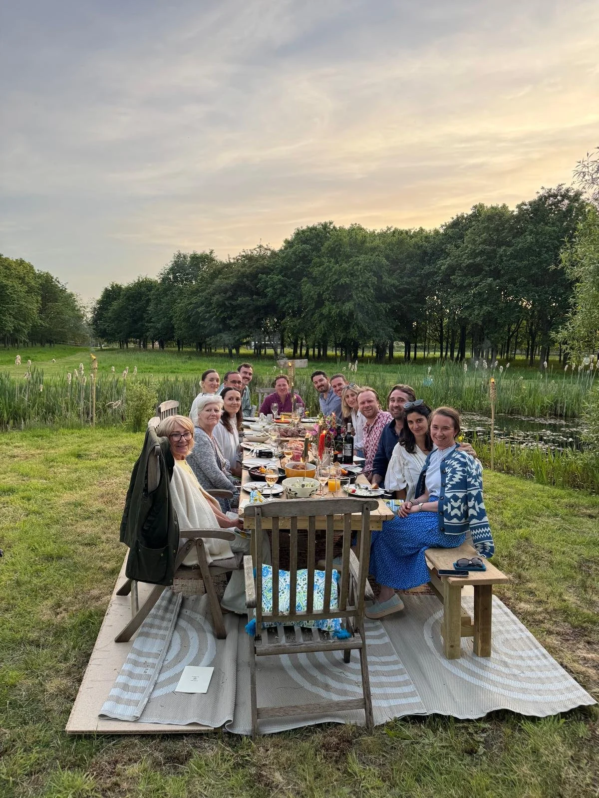 A group of people enjoying a meal at an outdoor dining table during sunset near a pond and trees.