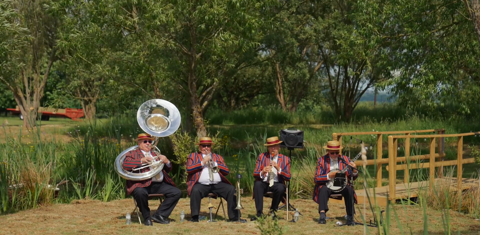 Four musicians dressed in matching striped blazers and straw hats, playing instruments outdoors: a tuba, a trumpet, a clarinet, and a guitar, seated on chairs in a grassy area with trees and a wooden platform in the background.