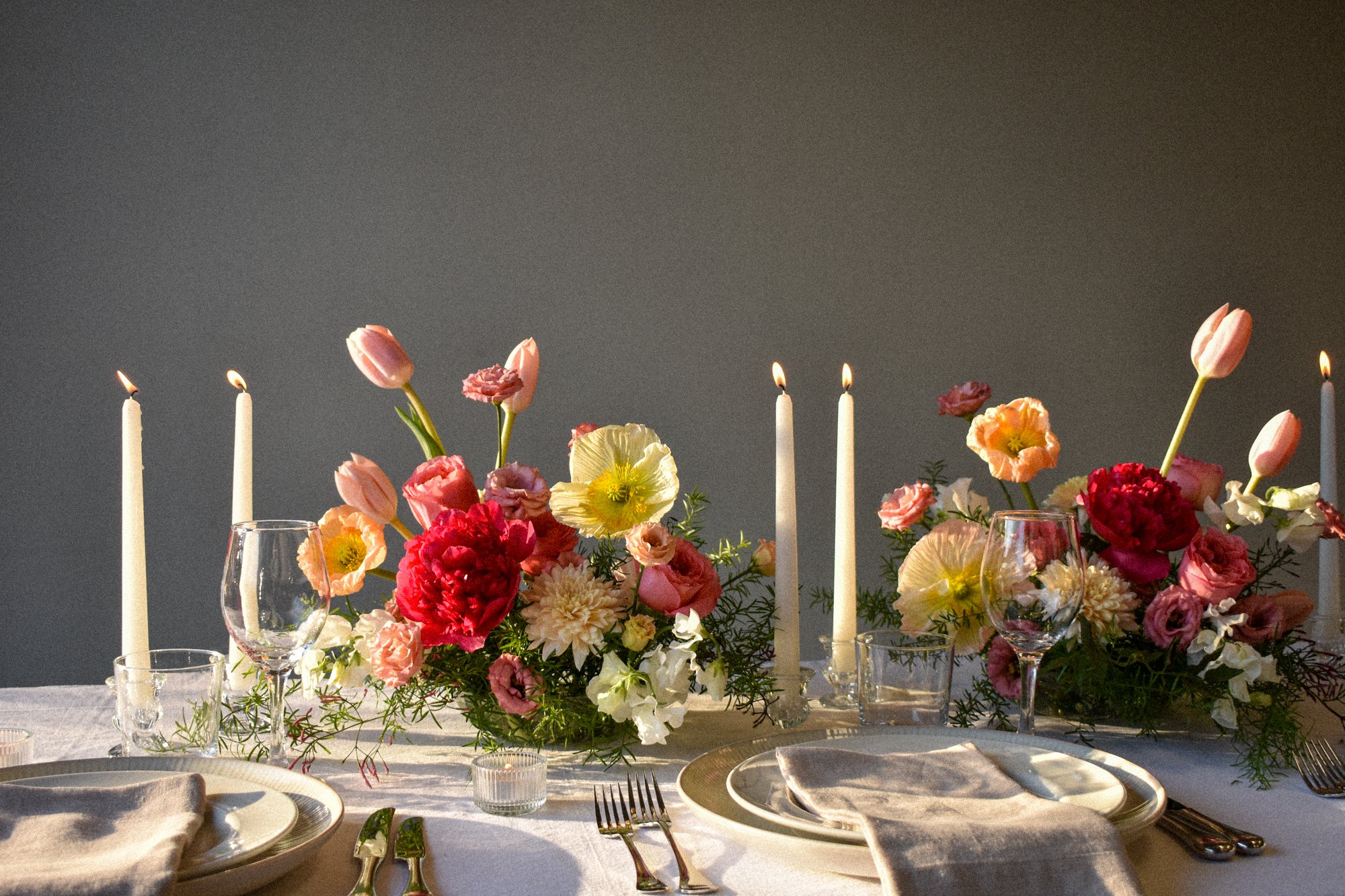 A dinner table setup with a floral centerpiece featuring pink, red, and yellow flowers, surrounded by candles, wine glasses, and plates, against a gray wall background.