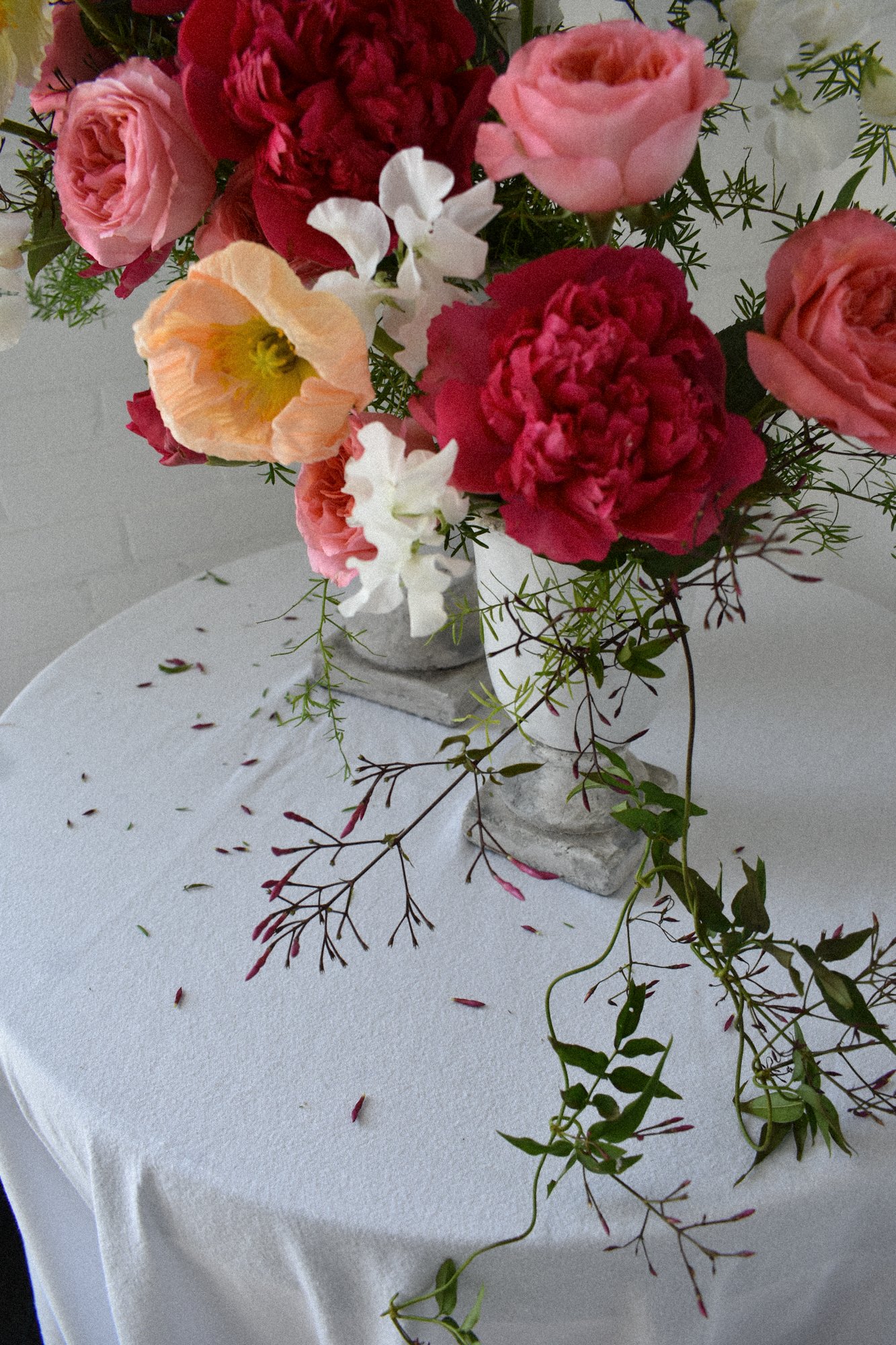 A floral arrangement in a white urn on a white round table, featuring pink garden roses, hot pink peonies, white sweet peas, poppies, flowering jasmine, and green foliage, with some fallen flower petals on the table.