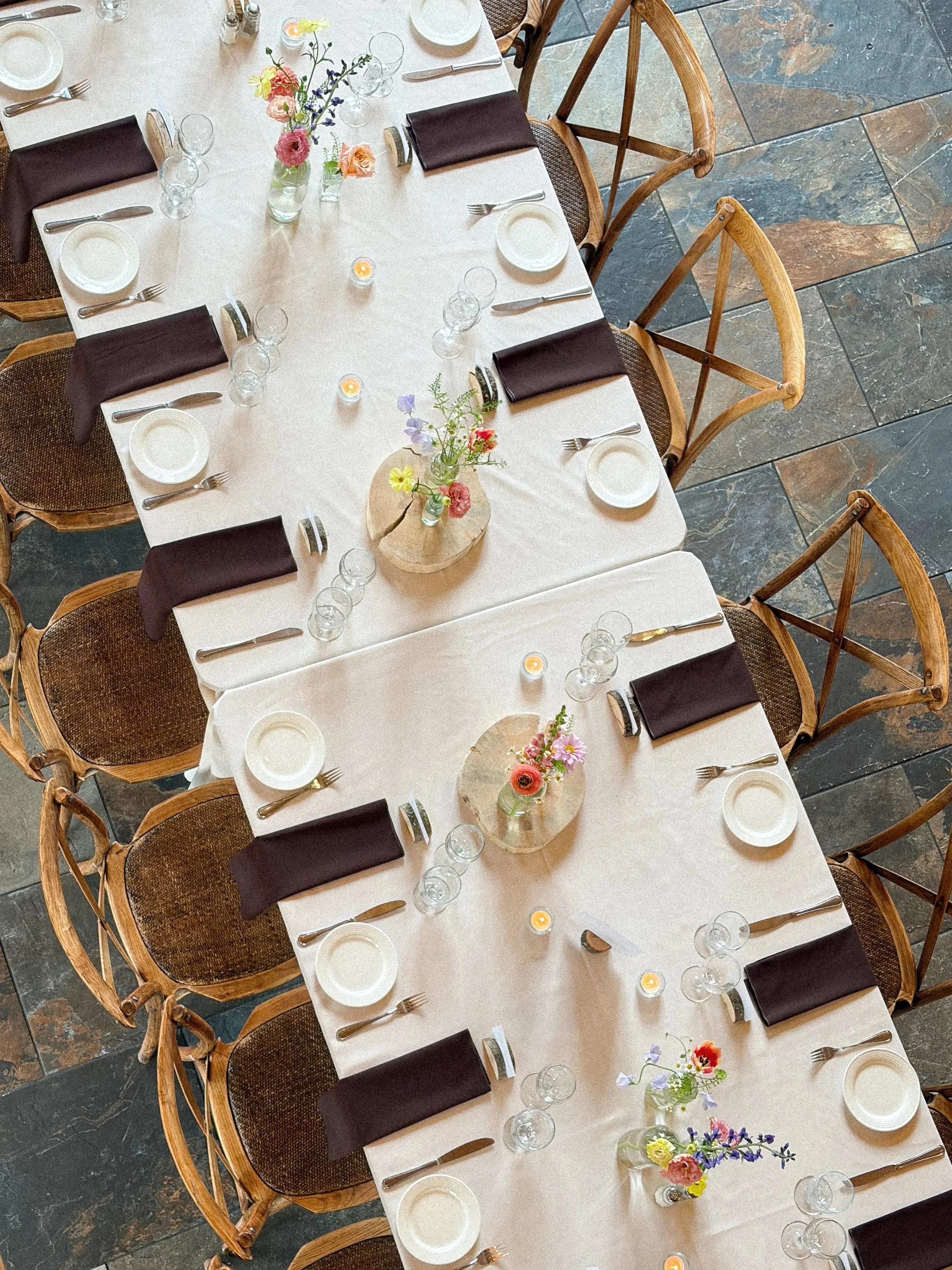 A top-down view of a long rectangular dining table set for a meal with white tablecloths, black napkins, white plates, silverware, wine glasses, small candles, and floral centerpieces.