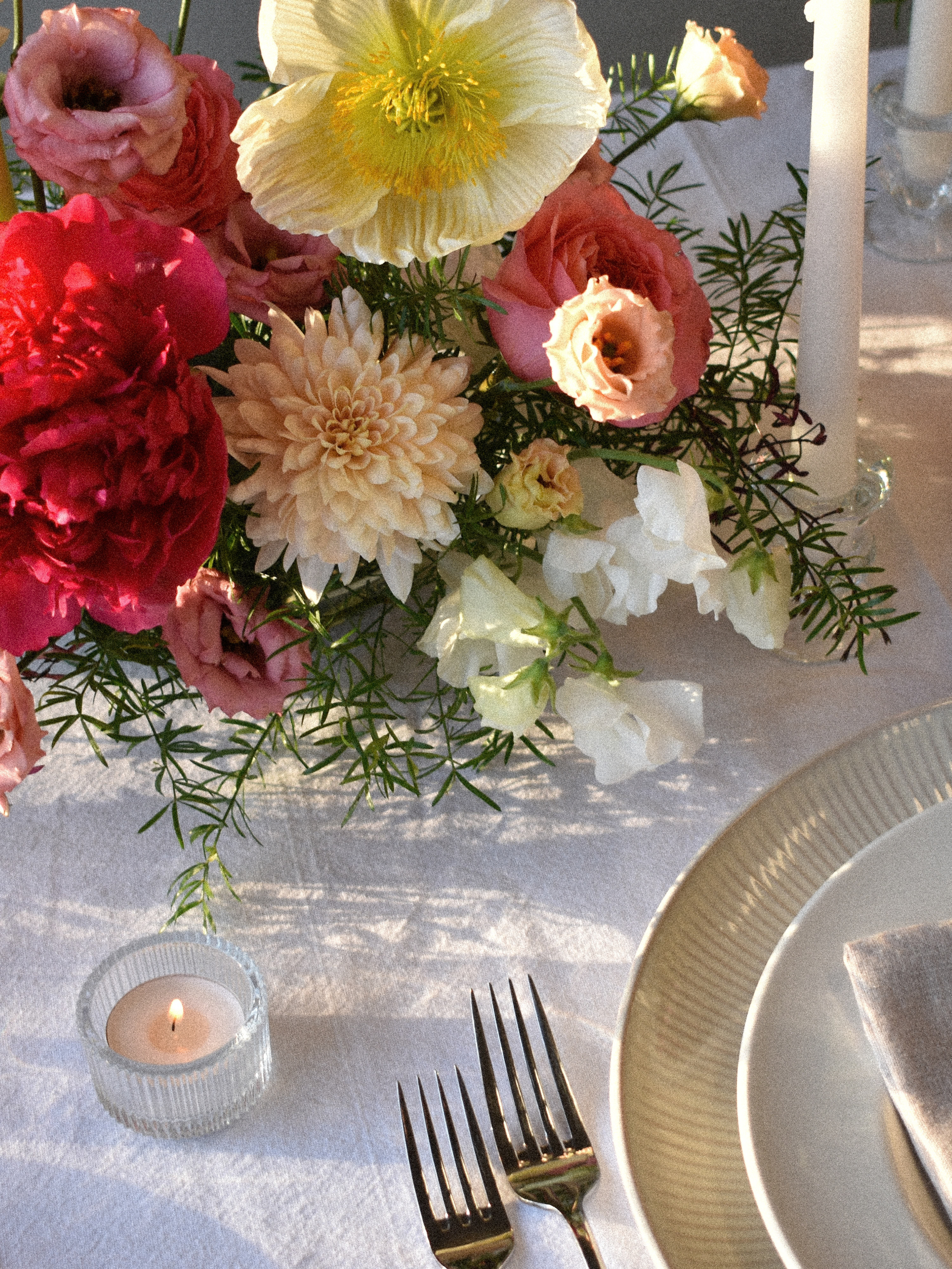 A floral centerpiece with pink, white, and peach flowers on a table set with white plates, forks, and a lit candle in a glass holder.