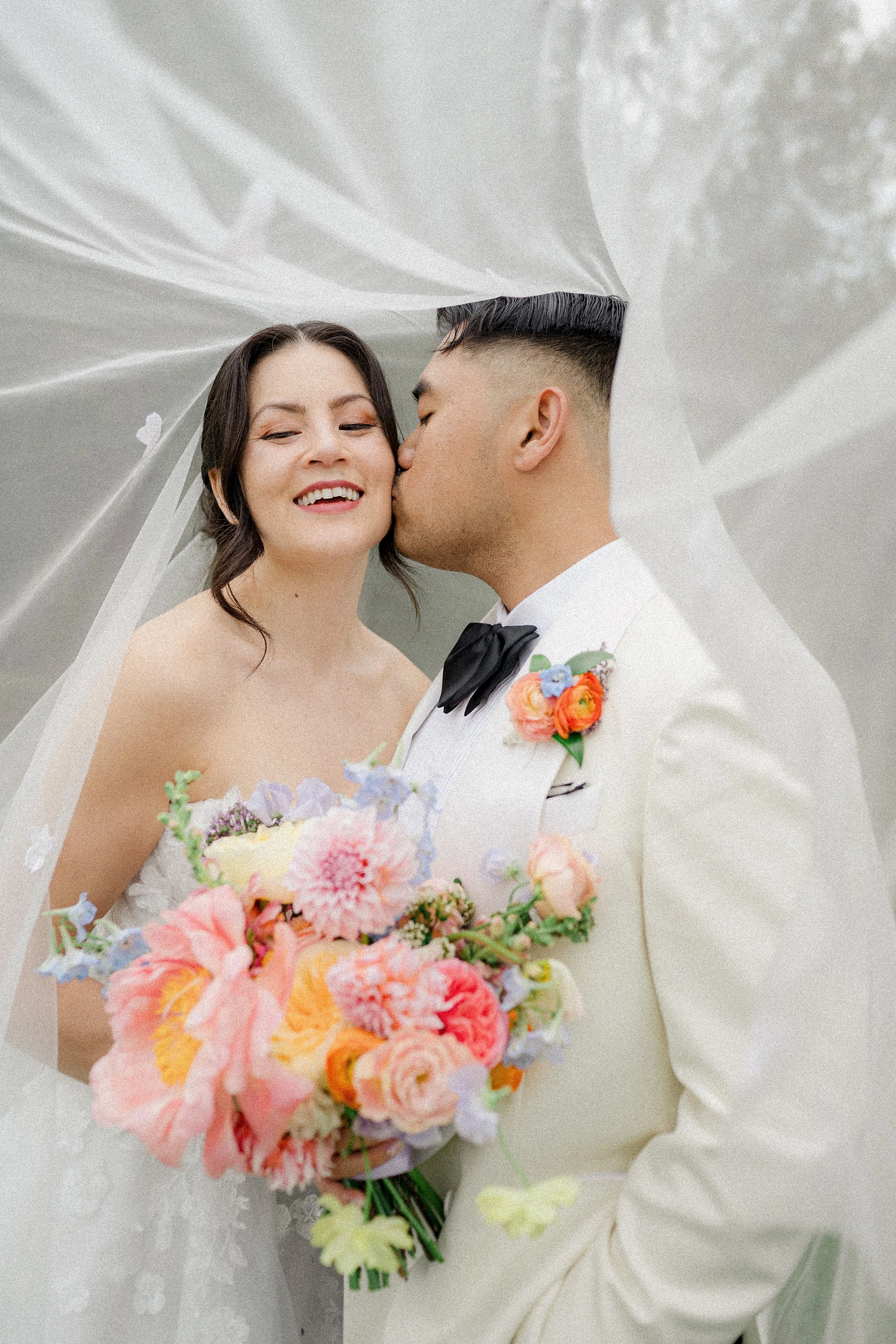 A bride and groom sharing a kiss under a white veil, with the bride holding a colorful bouquet of flowers, including pink, orange, and purple blooms.