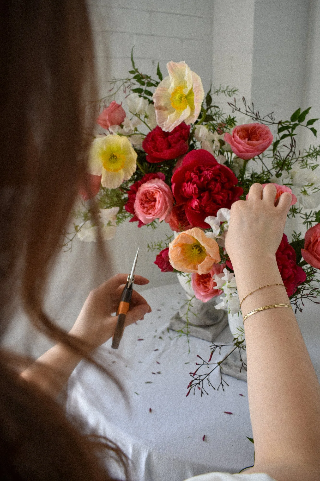 A person arranging a bouquet of pink, red, white, and yellow flowers on a white surface, using scissors to trim the stems.