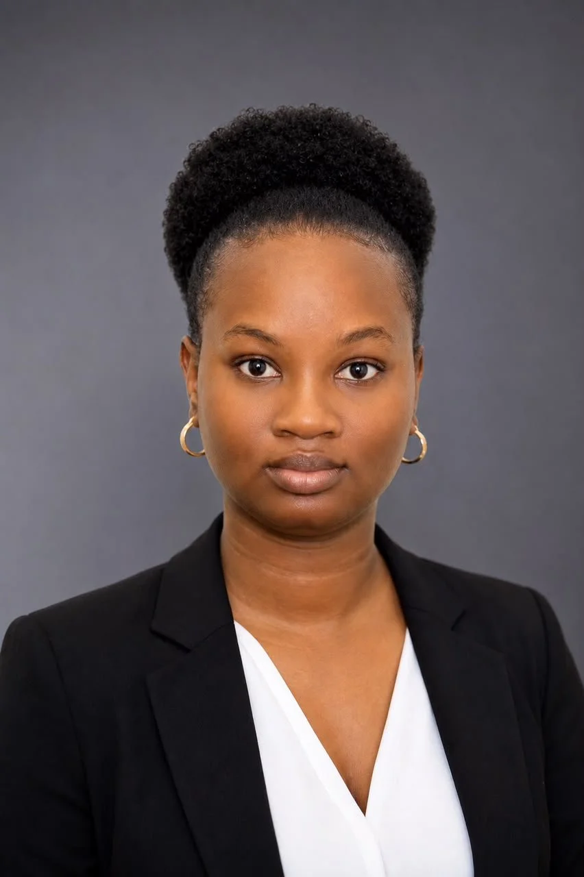 A professional portrait of a Black woman with short curly hair in a puff, wearing a black blazer over a white top, with hoop earrings, looking directly at the camera against a neutral gray background.