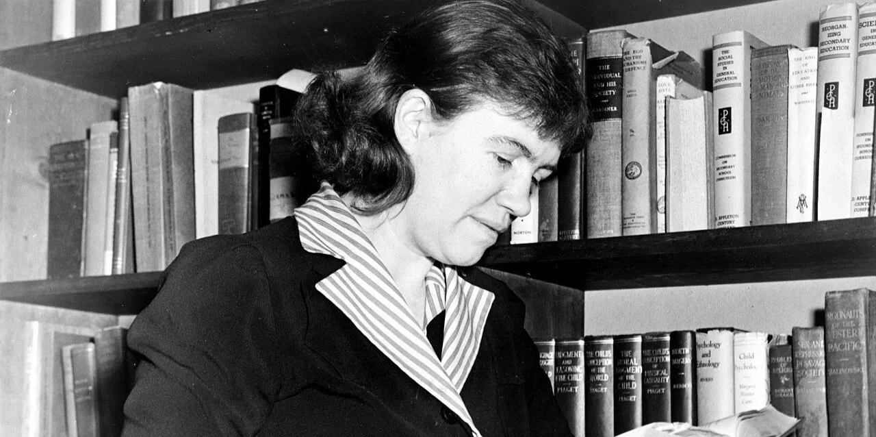 A woman with dark hair looking down at a book in front of a bookshelf filled with textbooks.