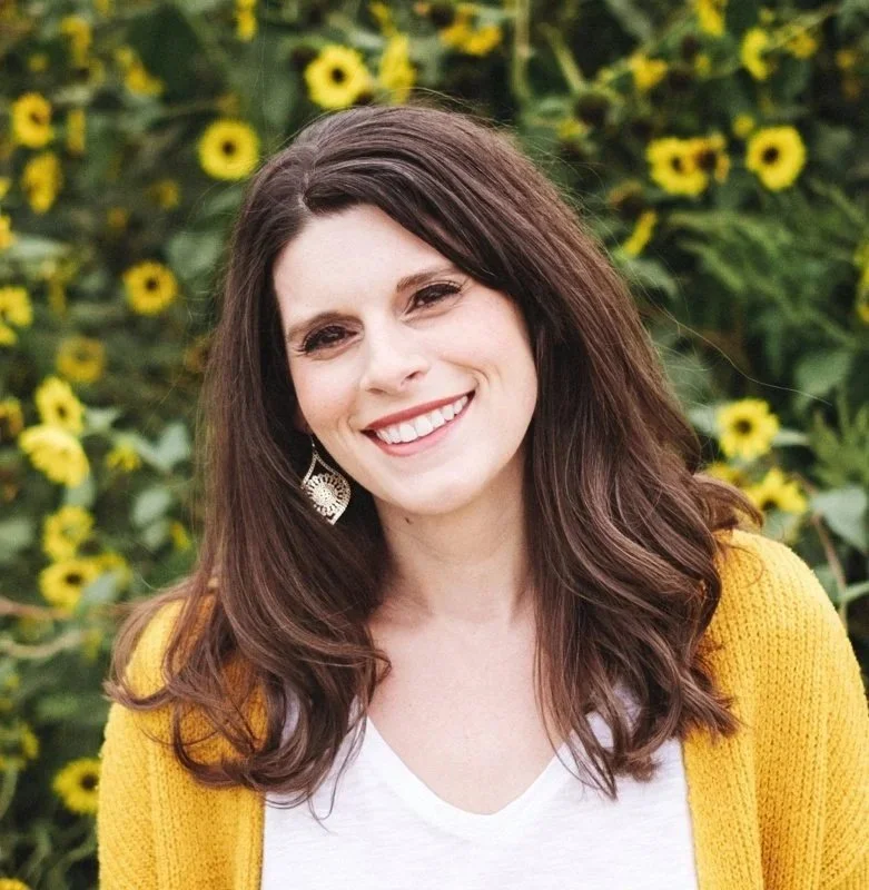 A woman with brown hair smiling outdoors in front of a sunflower field, wearing a yellow cardigan, white top, and earrings.