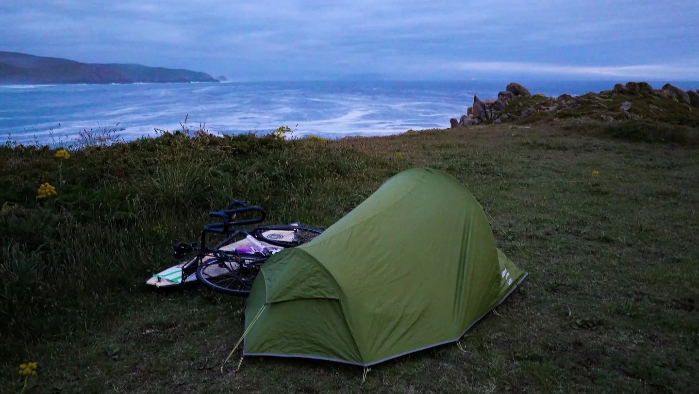 A green camping tent set up on grassy terrain near the coast with a bicycle lying on the ground beside it, overlooking the ocean with waves and a distant landmass under a cloudy sky.