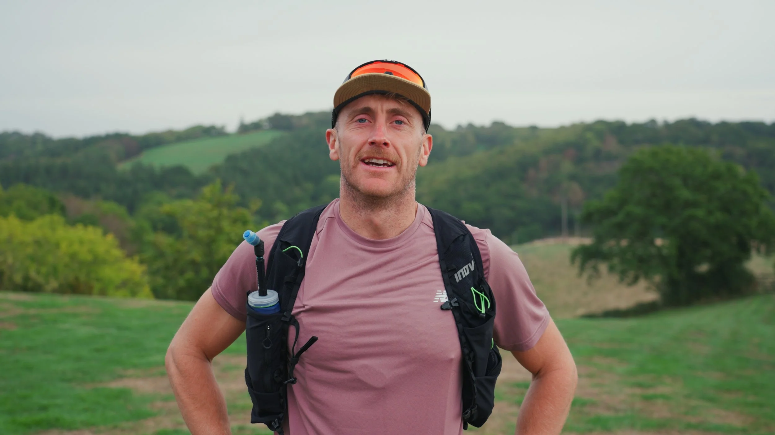 A man in a pinkish t-shirt with a backpack, cap, and sunglasses standing outdoors in a green landscape with hills and trees at a trail running retreat in Dartmoor.